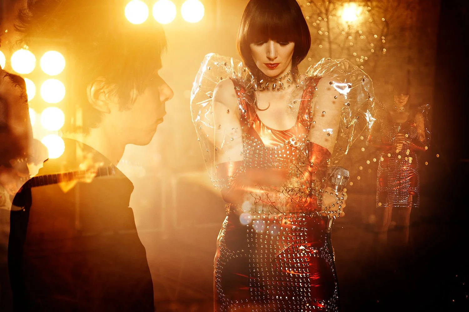 Karen O of the Yeah Yeah Yeah's in a shiny, metallic red dress with clear, puffy sleeves and arm bands, holding a microphone and standing in front of bright, warm stage lights, with a man in a dark outfit 