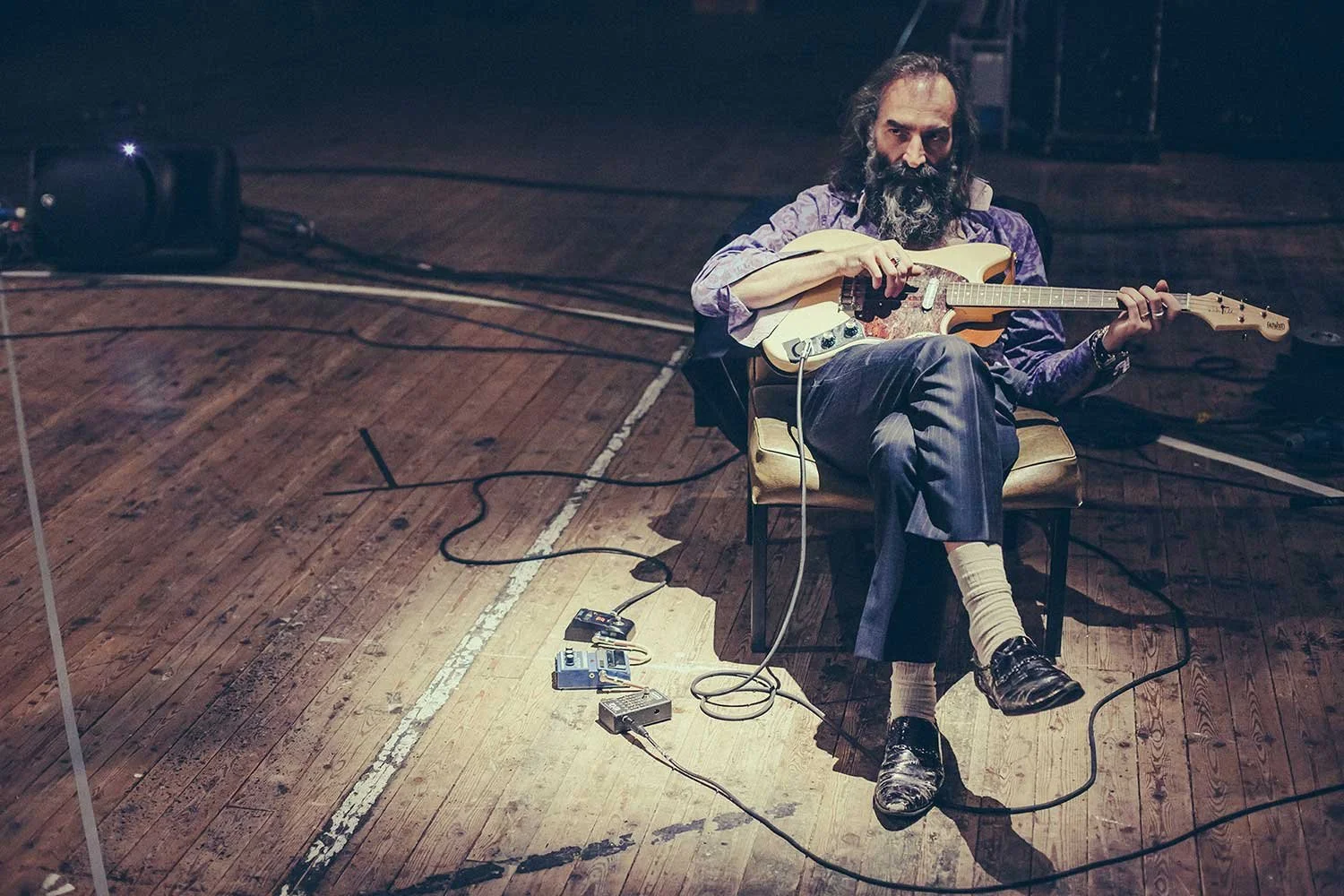 Warren Ellis with long hair and beard, wearing a purple shirt and striped pants, is sitting on a chair on a wooden stage, playing an electric guitar. He is surrounded by guitar pedals and cables on the stage.