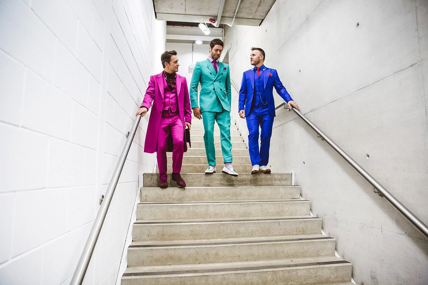 Gary Barlow, Mark Owen and Howard Donald of Take That dressed in brightly colored suits walking down stairs in a concrete and white-walled underground corridor.