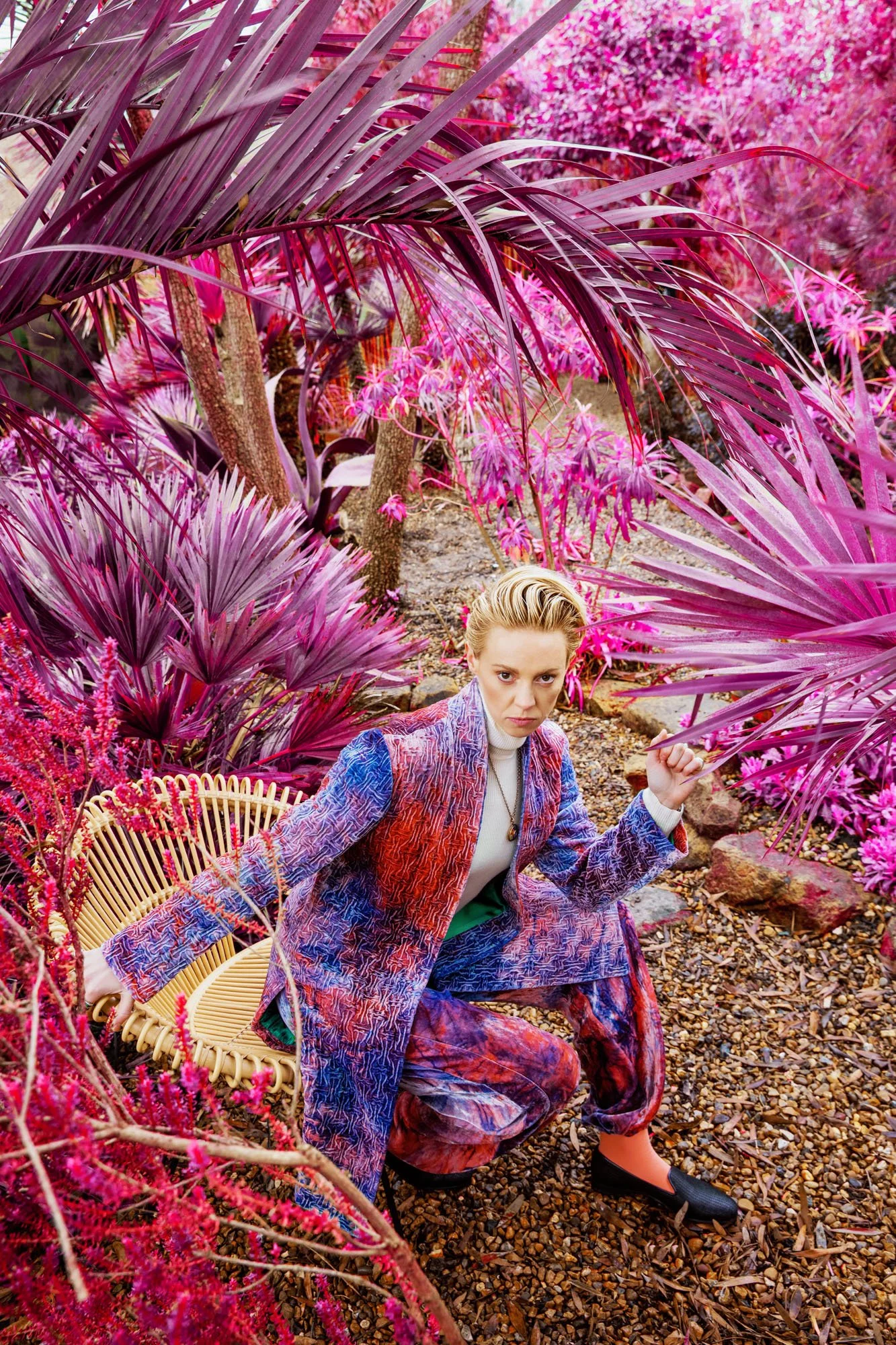 La Roux with blonde hair styled back, wearing a colorful patterned blazer and matching pants, crouches in a lush garden of pink and purple plants and flowers, holding a branch in each hand.