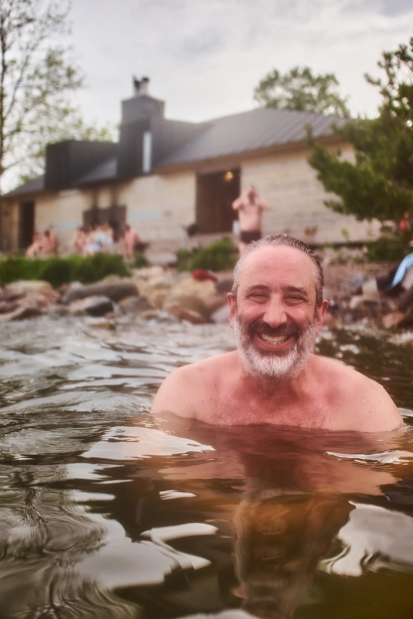 A happy David Baddiel with a grey beard swimming in a natural hot spring or river near a stone building with other people relaxing nearby.