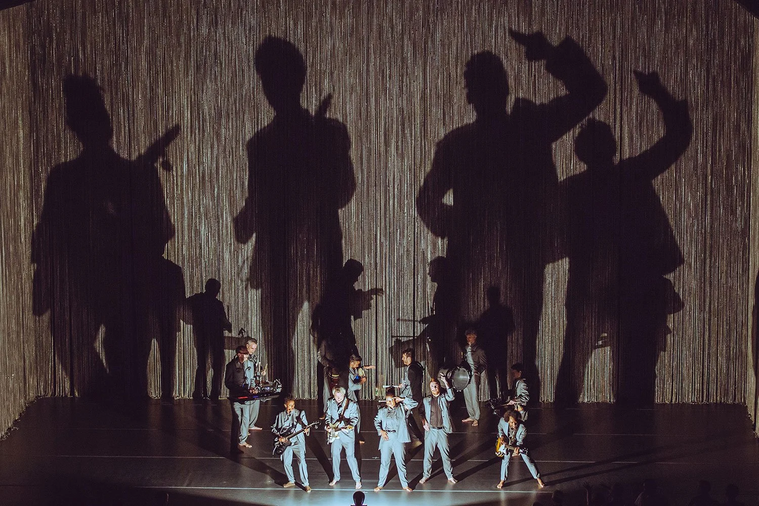 David Byrne live. A group of performers in matching grey suits on stage, with their large shadow silhouettes projected on a curtain behind them, creating a dramatic visual effect.
