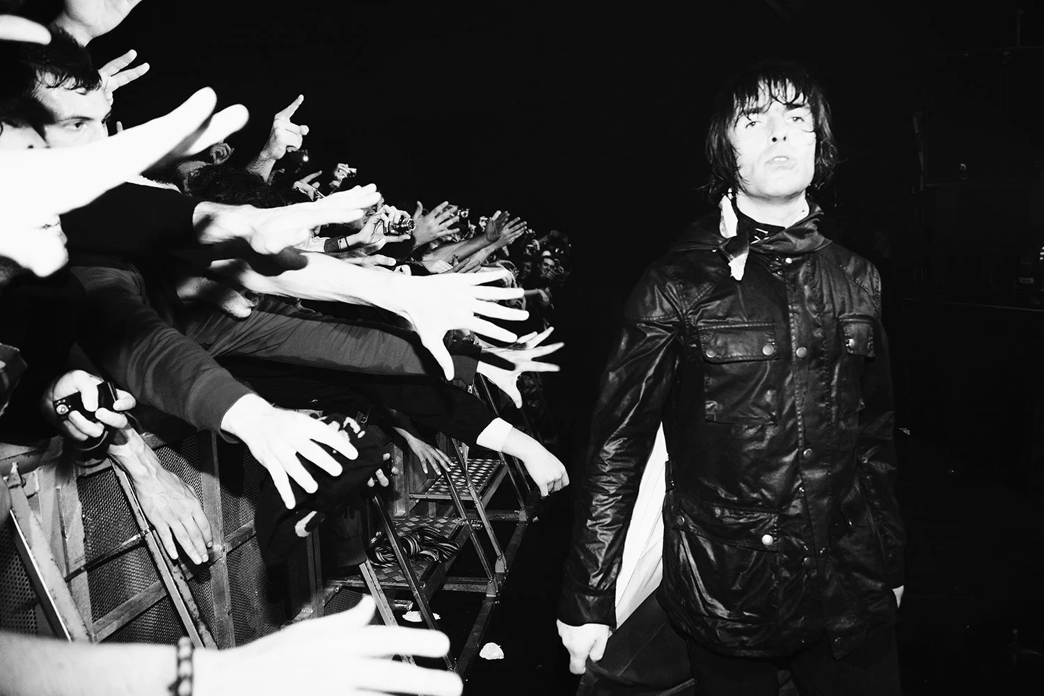 Liam Gallagher with shoulder-length hair wearing a black leather jacket walking past a crowd of fans reaching out to him.