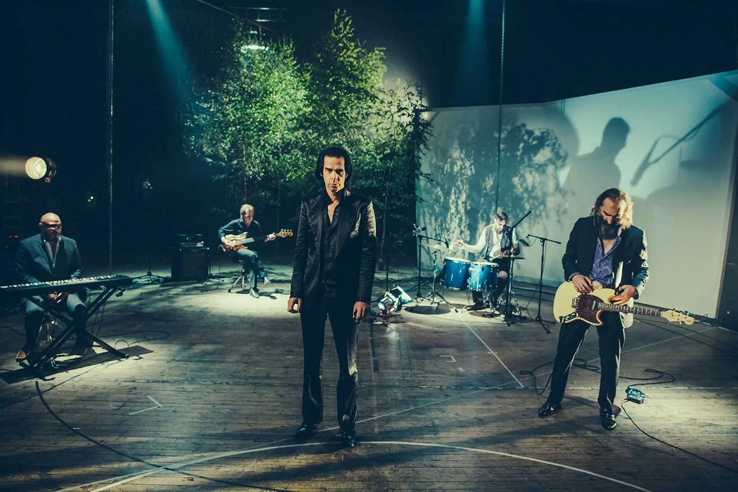 Nick Cave and the Bad Seeds performing on stage with a scenic backdrop of trees and shadows of leaves, featuring Nick cave, Warren Ellis, a drummer, and a keyboard player.