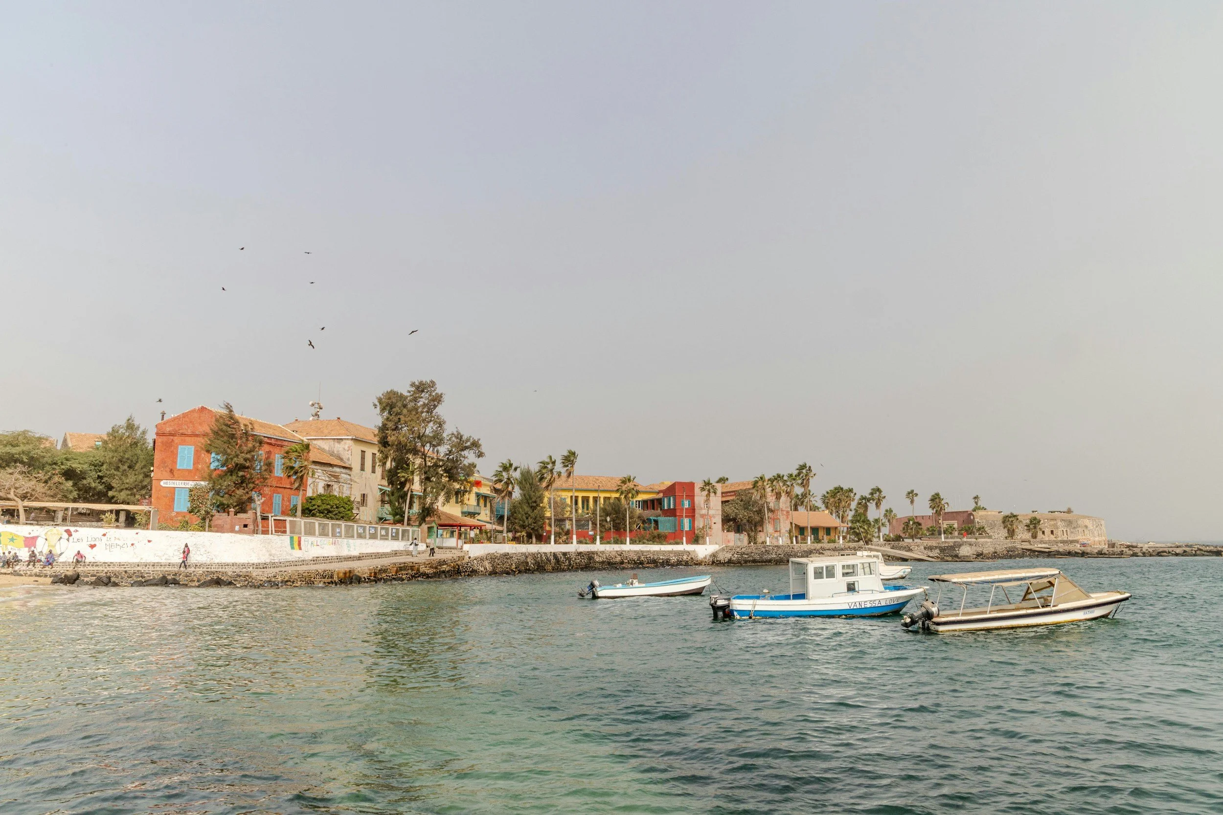 View of a seaside town with colorful buildings, palm trees, and boats docked in the water under a cloudy sky.
