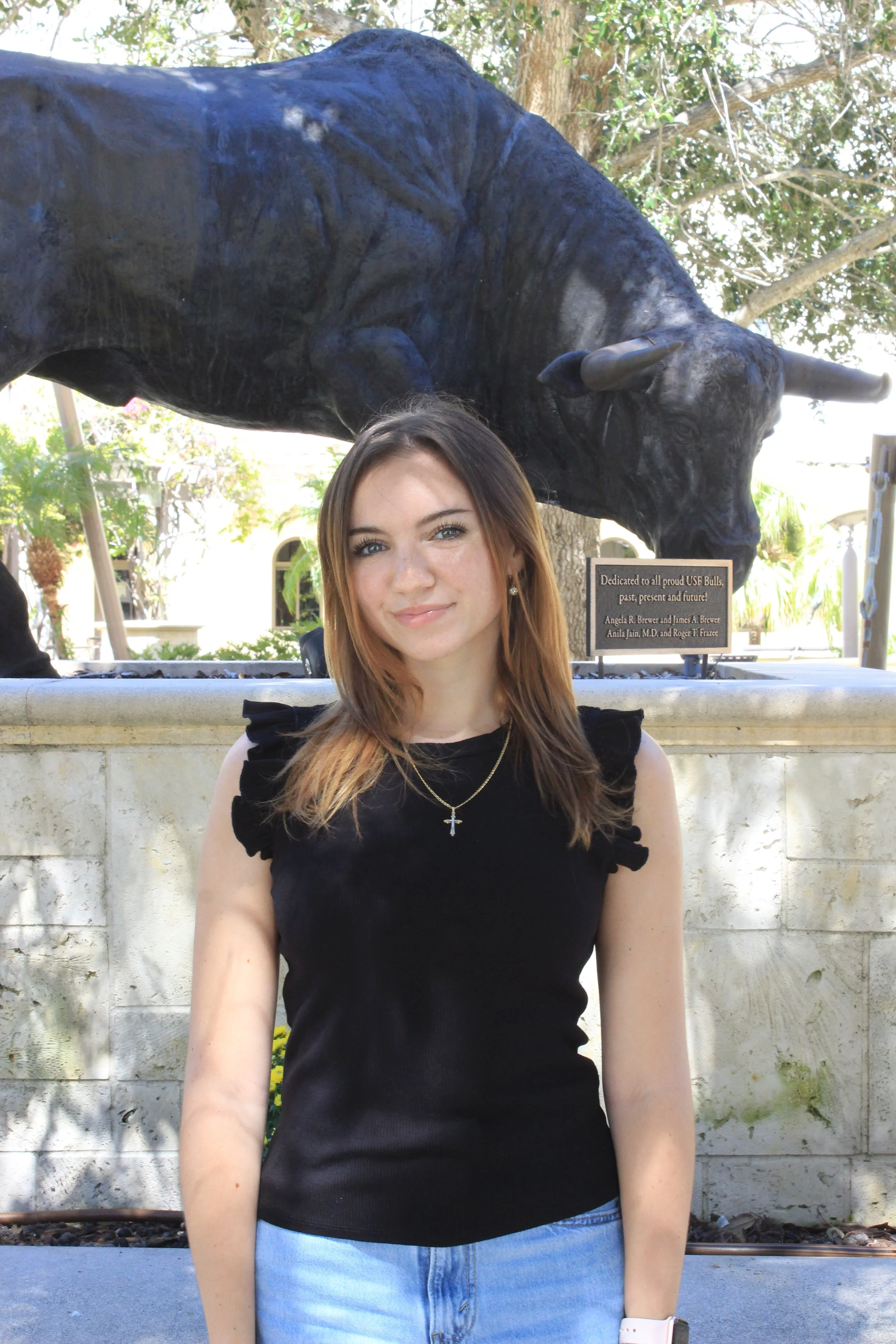 Young woman with long brown hair, wearing a black sleeveless top and jeans, standing in front of a large bull statue in a park or outdoor area.