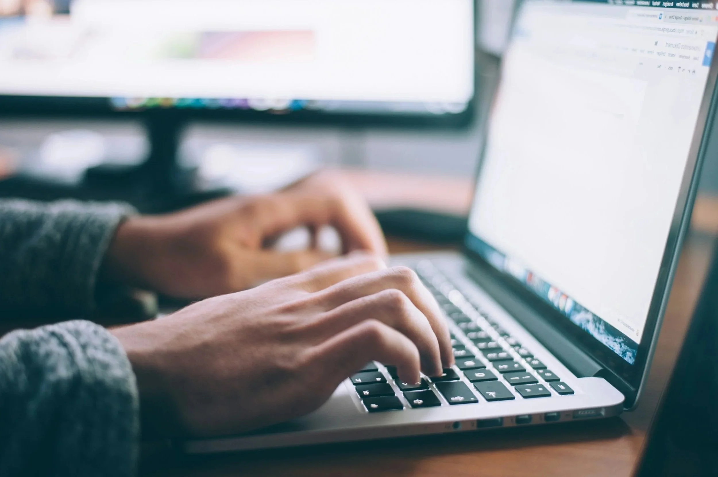 Close-up of hands typing on a laptop keyboard with a blurred computer monitor in the background.