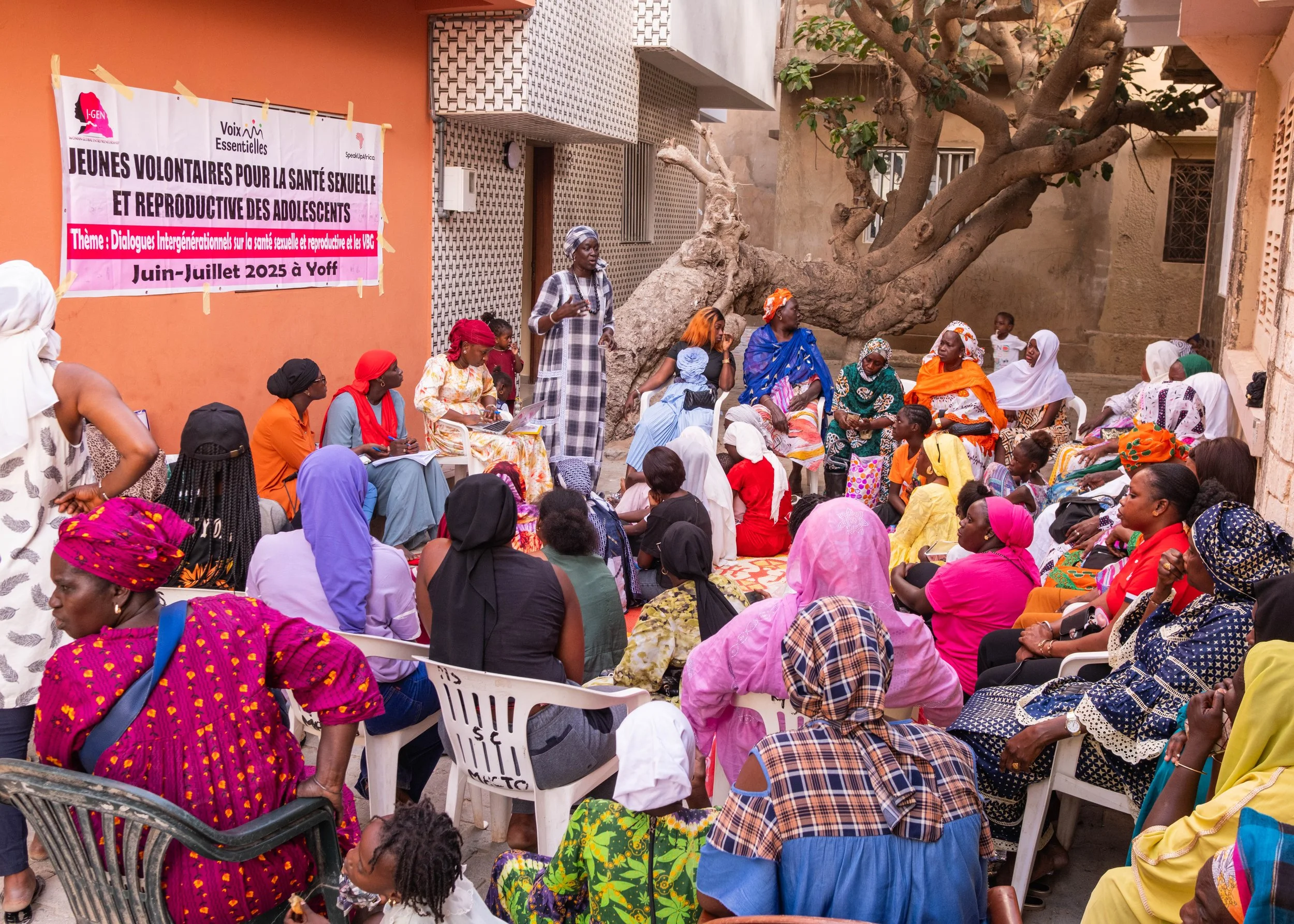 A group of women and children seated outdoors, listening to a speaker during a community event. The setting appears to be in an urban area with colorful walls, a large tree, and various seating arrangements. A banner in the background indicates the event is related to sexual and reproductive health education for adolescents in Yoff, June-July 2025.