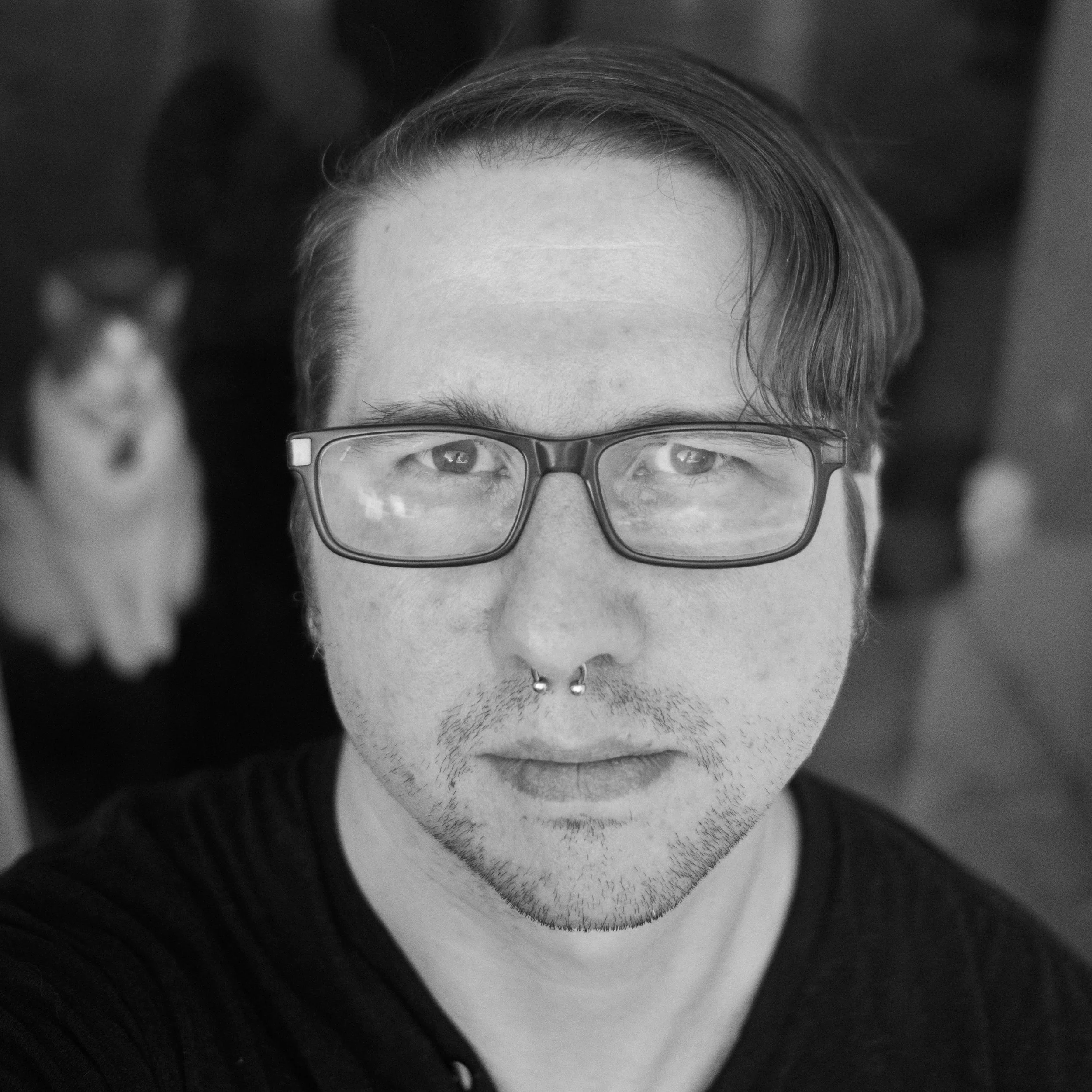 A black and white close-up photo of a man with glasses and a septum piercing, looking directly at the camera, with a blurred cat in the background.