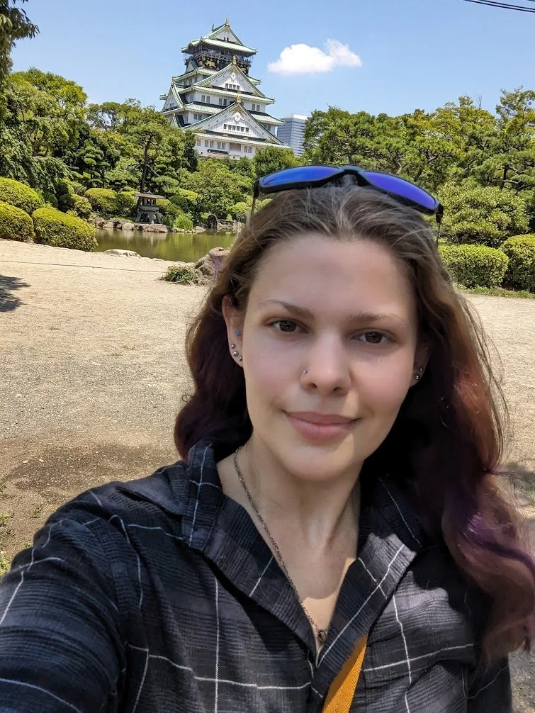 A young woman with long hair, sunglasses on her head, and multiple earrings taking a selfie in a traditional Japanese garden with a pond and the Himeji Castle in the background.
