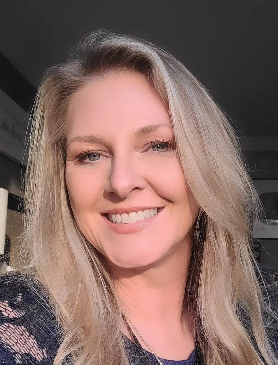 A smiling woman with long blonde hair, blue eyes, and fair skin, indoors with a dark background.