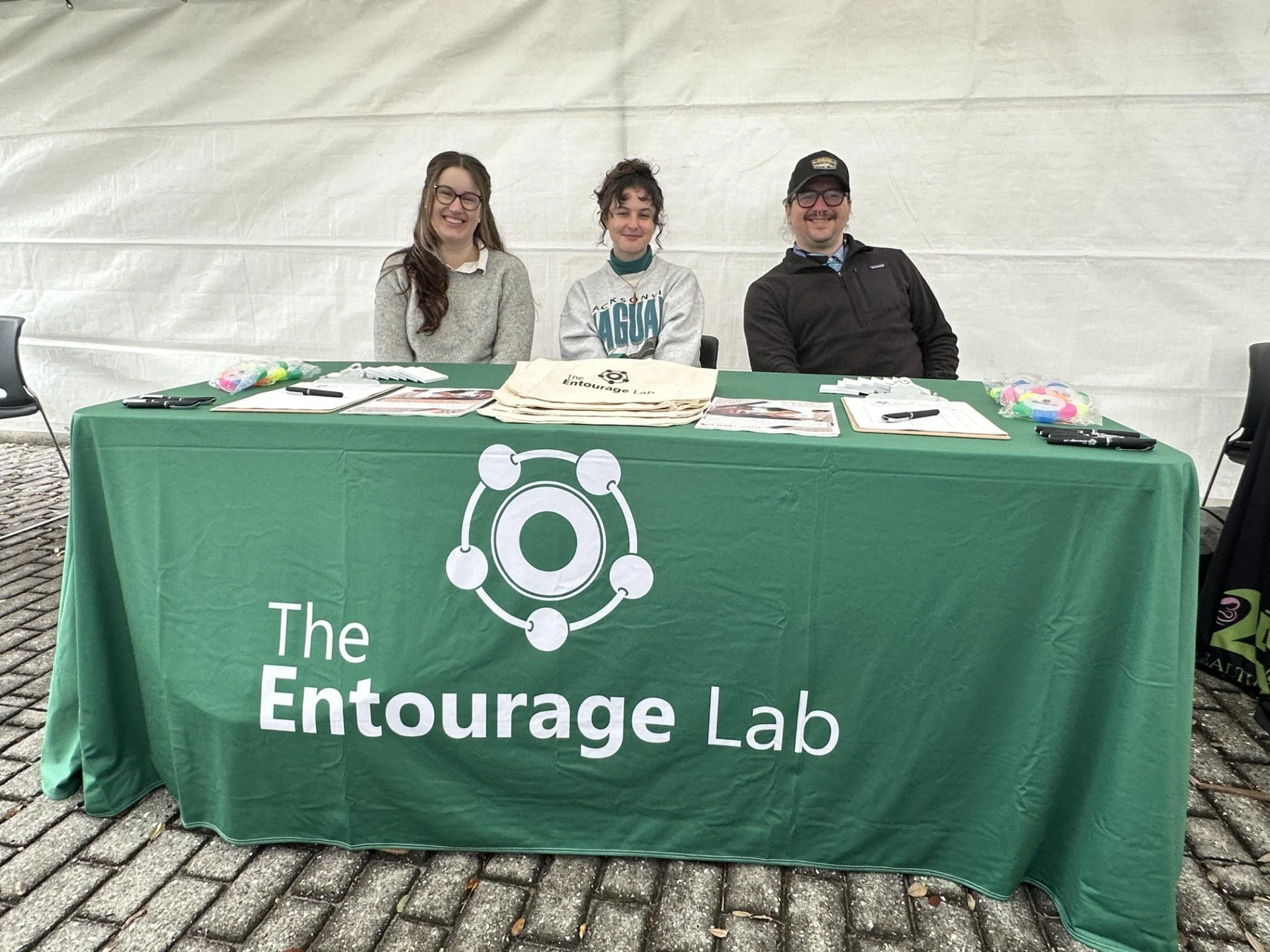 Three people sitting behind a green tablecloth with 'The Entourage Lab' logo and name, at an event booth with papers and supplies on the table.