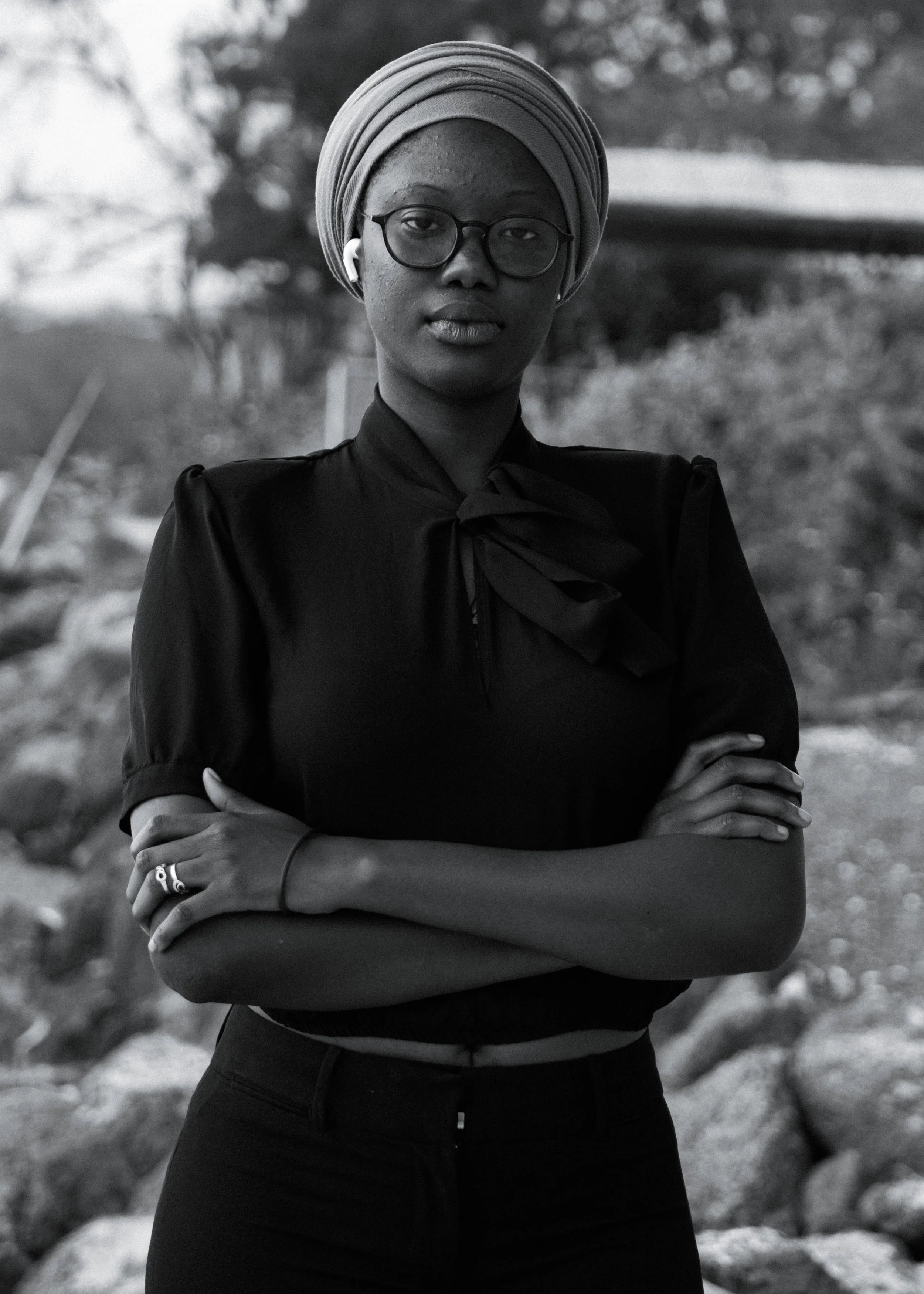 A woman with glasses and a headwrap standing outdoors with arms crossed.