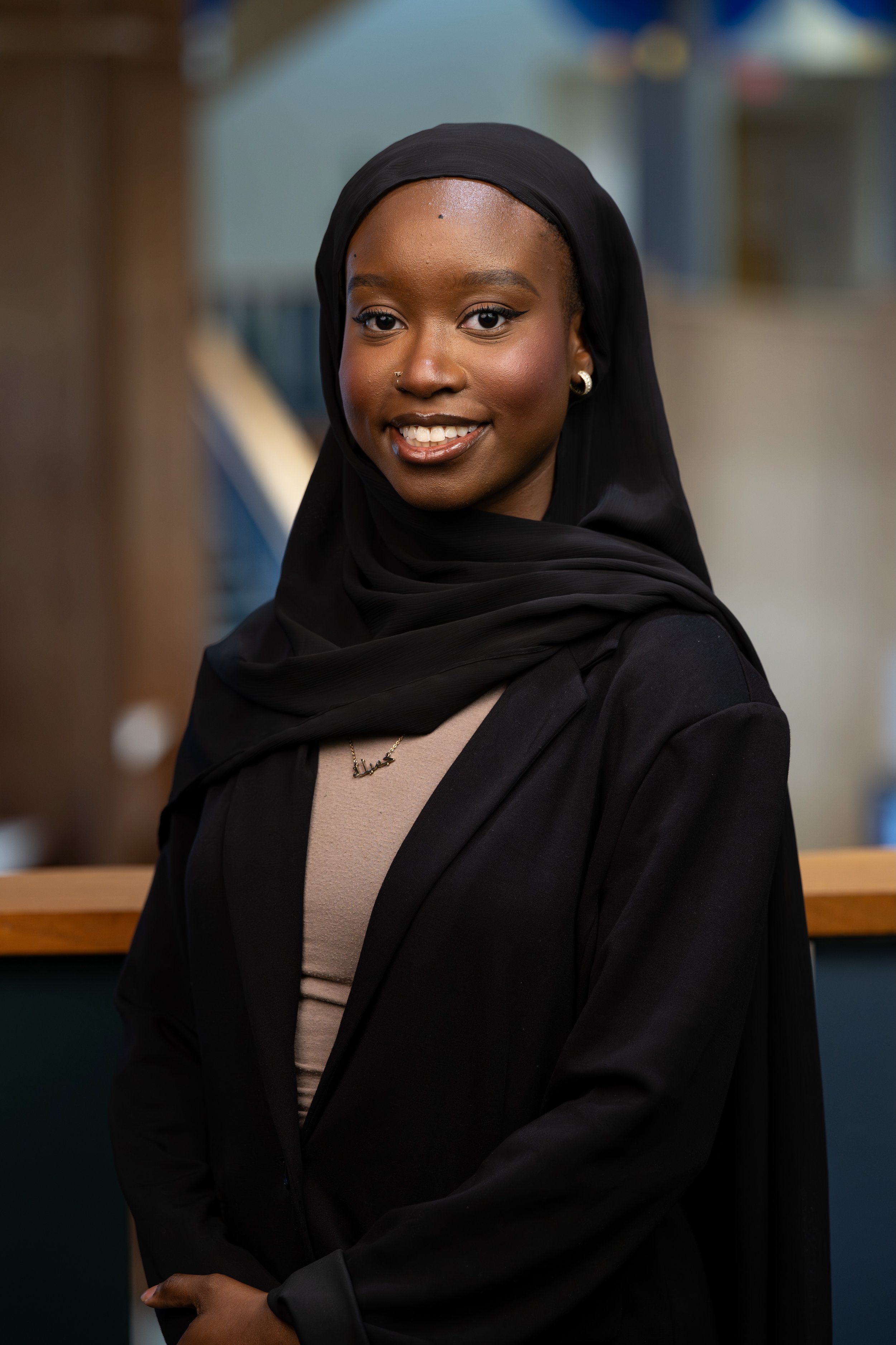 A smiling young woman with dark skin, wearing a black headscarf, a black blazer, and a beige top, standing in an indoor setting.