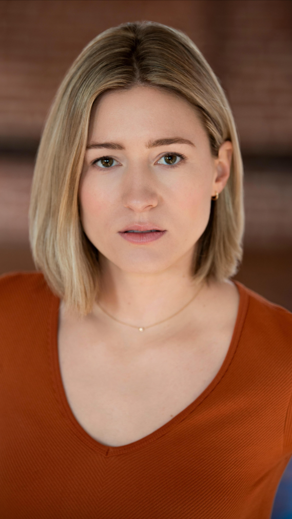 A woman with shoulder-length blonde hair and fair skin, wearing an orange top and a delicate gold necklace, looking directly at the camera with a neutral expression.