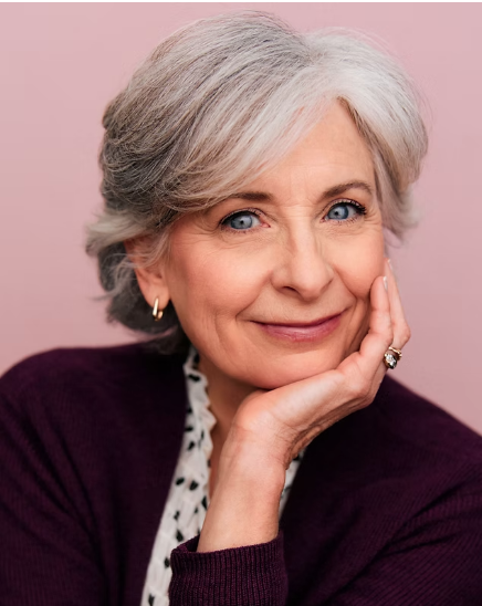 Close-up portrait of a smiling older woman with short, gray hair, wearing a burgundy sweater and gold earrings, resting her face on her hand against a pink background.