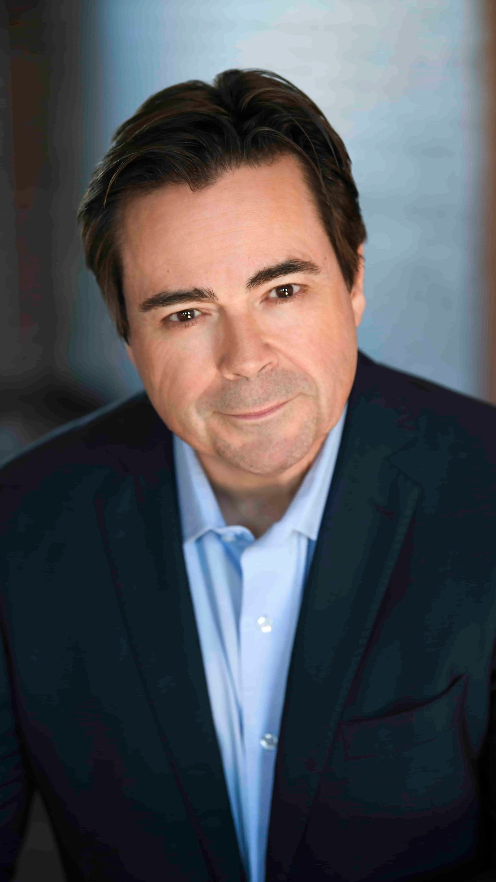 A professional headshot of a man in a dark suit and light blue shirt, smiling slightly, with dark brown hair and a blurred blue background.