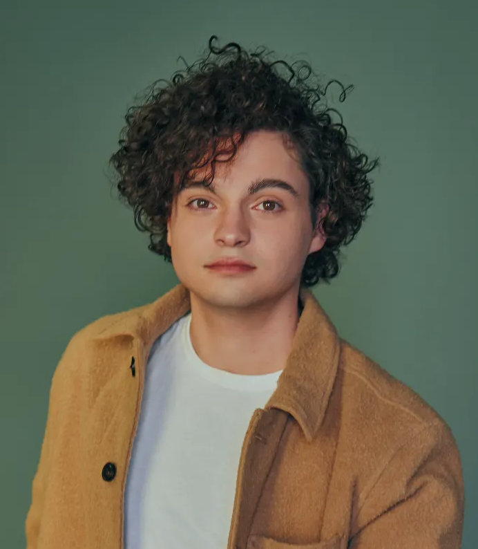 Portrait of a young man with curly dark hair, wearing a white t-shirt and a tan jacket, against a green background.