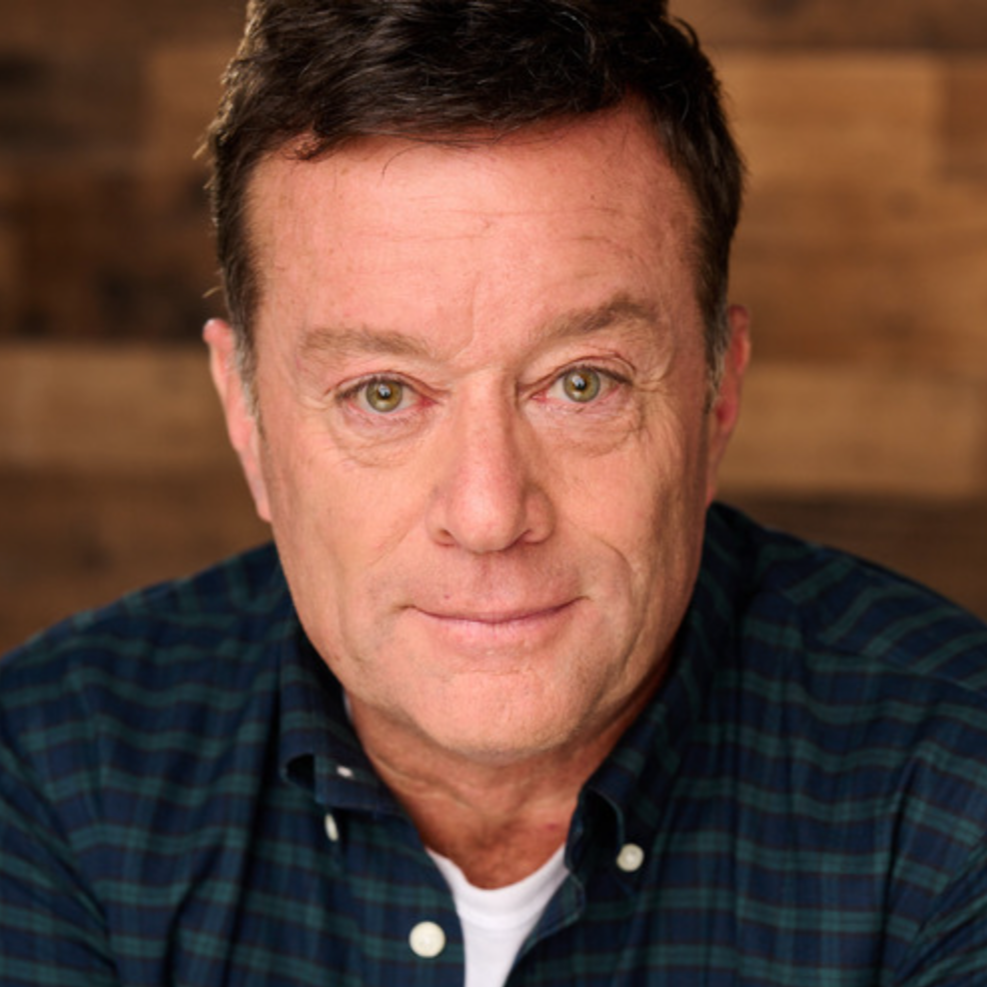 A middle-aged man with brown hair, green eyes, and a slight smile, wearing a blue plaid shirt, sitting in front of a wooden background.