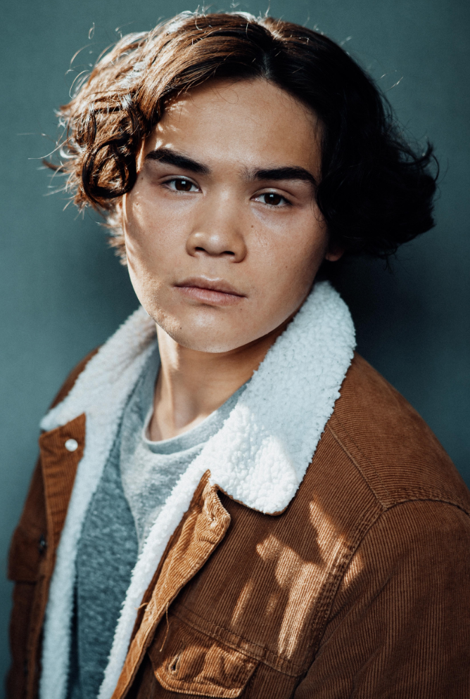 A young man with dark, wavy hair and tan skin, wearing a brown jacket with a white, fuzzy collar over a gray shirt, looking directly at the camera against a teal background.