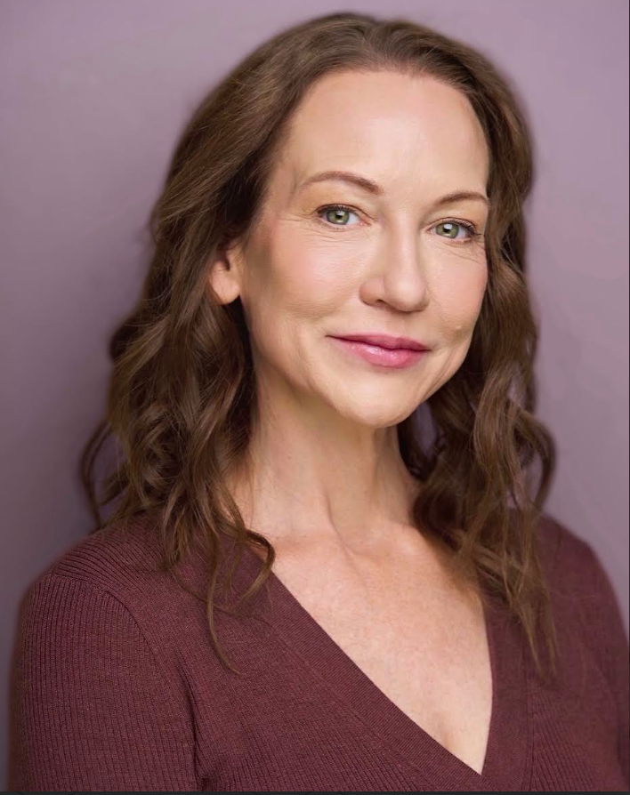 Portrait of a woman with shoulder-length wavy brown hair, light skin, green eyes, wearing a maroon V-neck top, against a purple background.