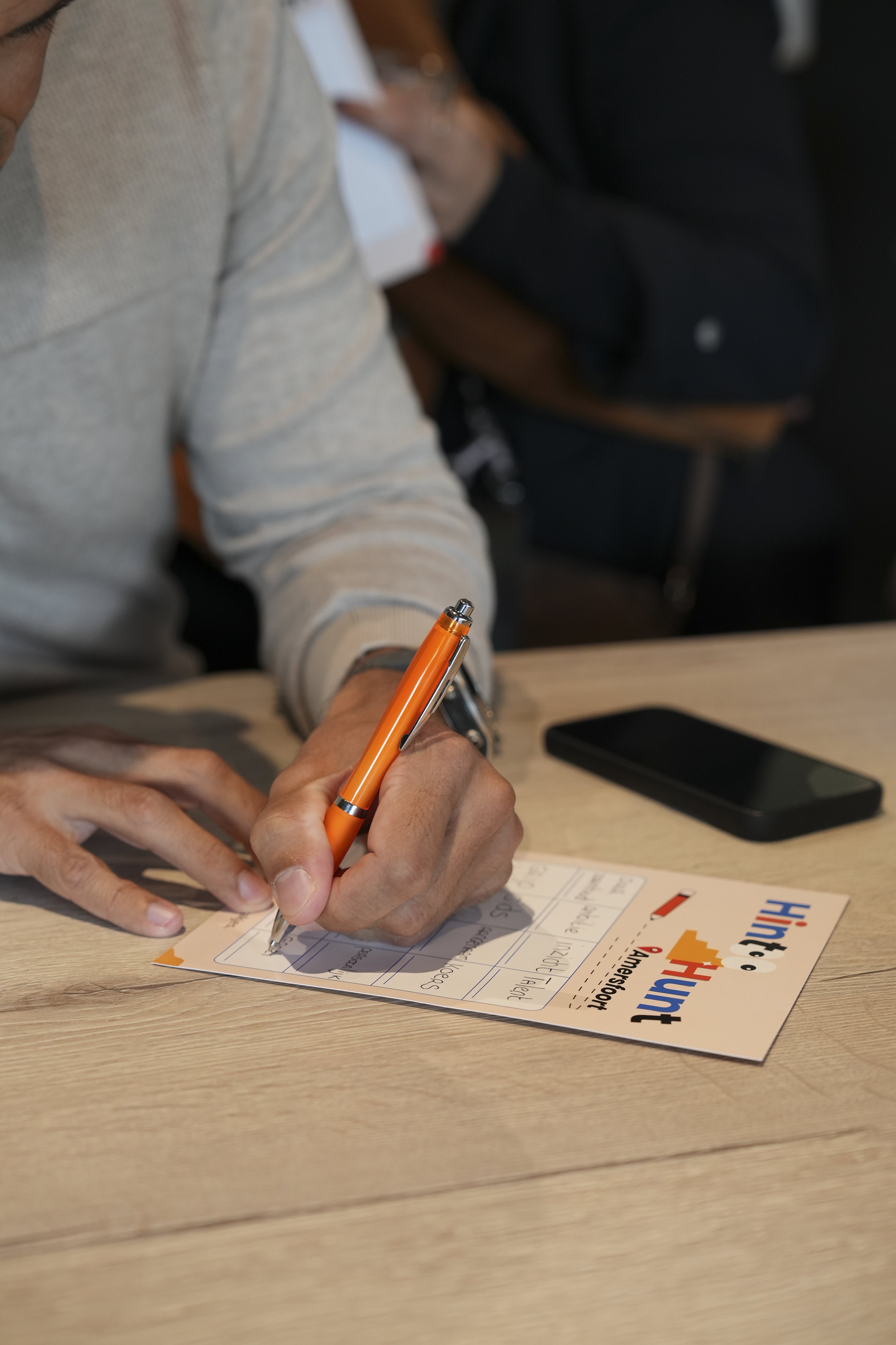Person signing a postcard with an orange pen at a table, with a smartphone beside the postcard.