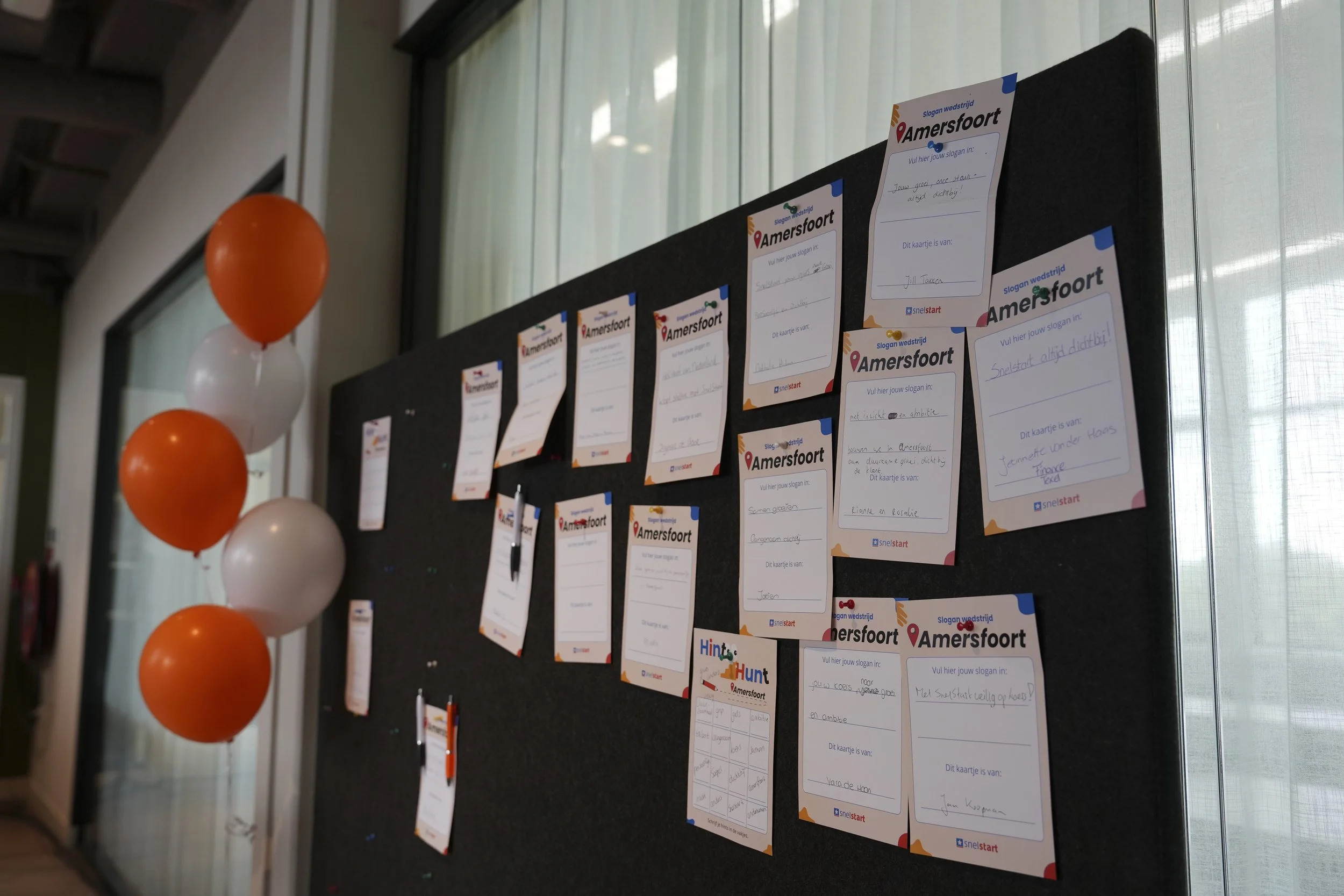 A black bulletin board with multiple handwritten notes and slogans for Amersfoort, decorated with orange and white balloons on the side, near a window with sheer curtains.