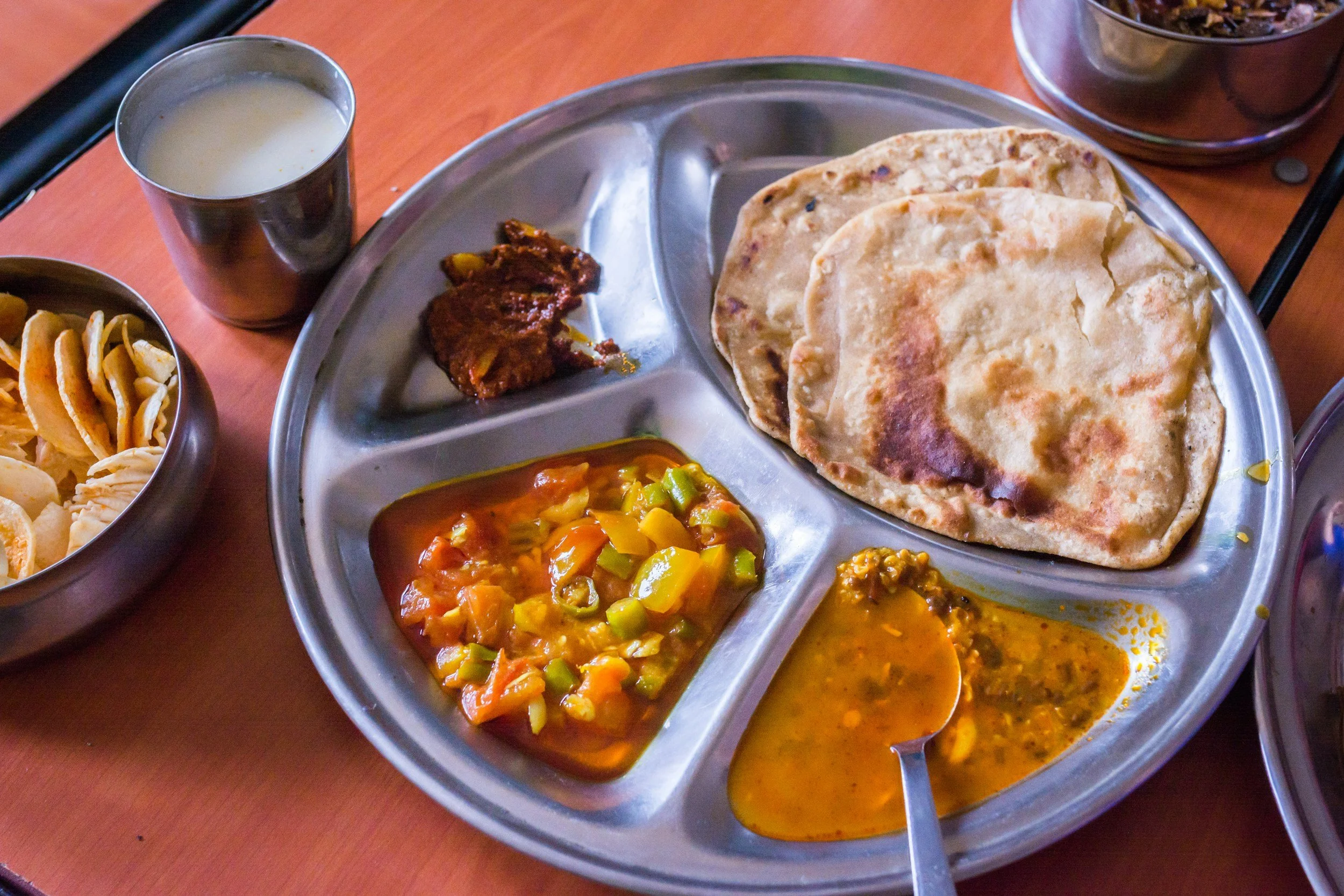 A metal tray with Indian meal items including chapatis, vegetable curry, lentils, and a side of chutney, with small bowls of yogurt and papadums nearby.