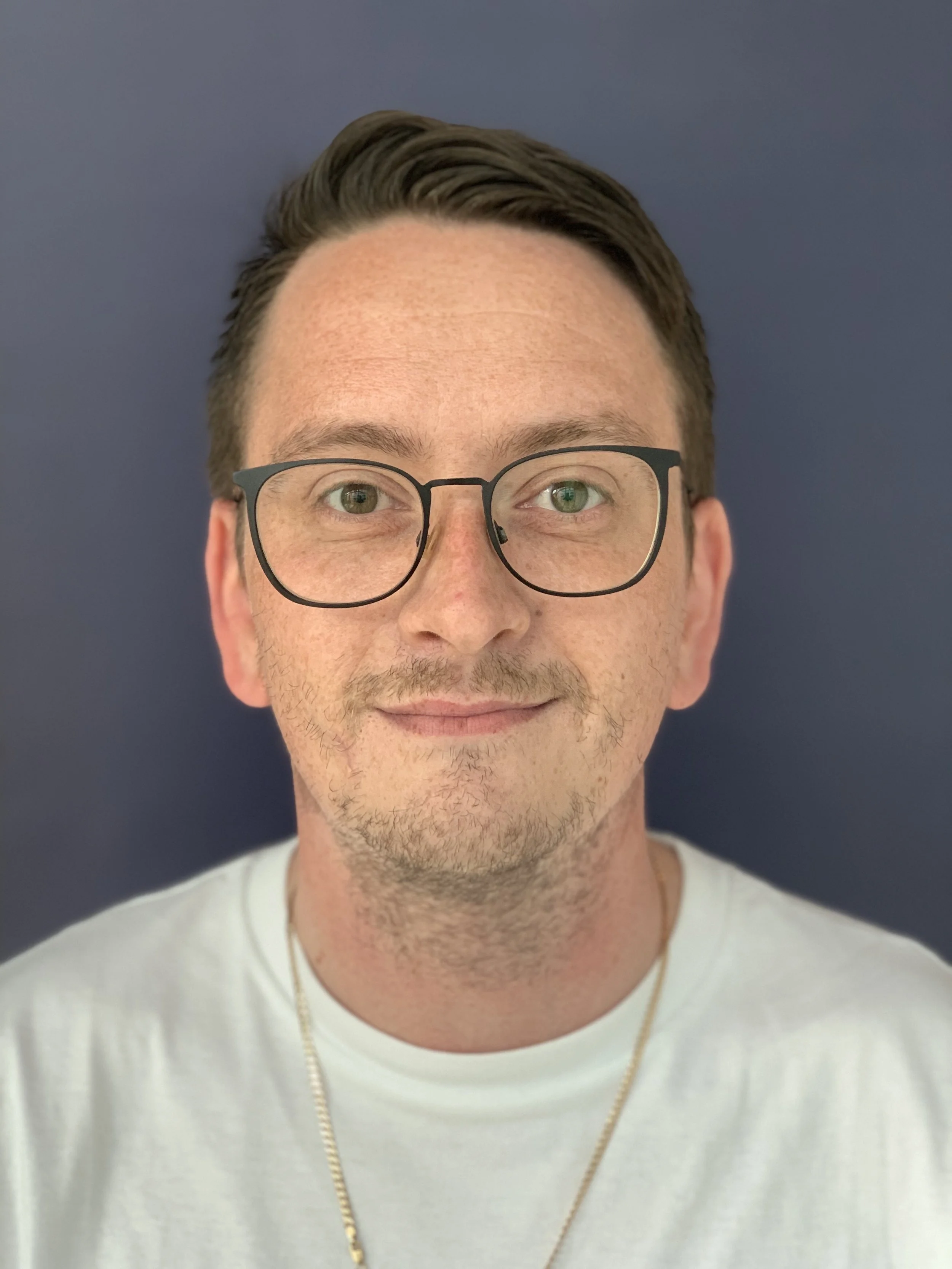 A man with short dark hair, glasses, light skin with freckles, near beard, smiling, wearing a white T-shirt and a gold chain, against a dark blue background.
