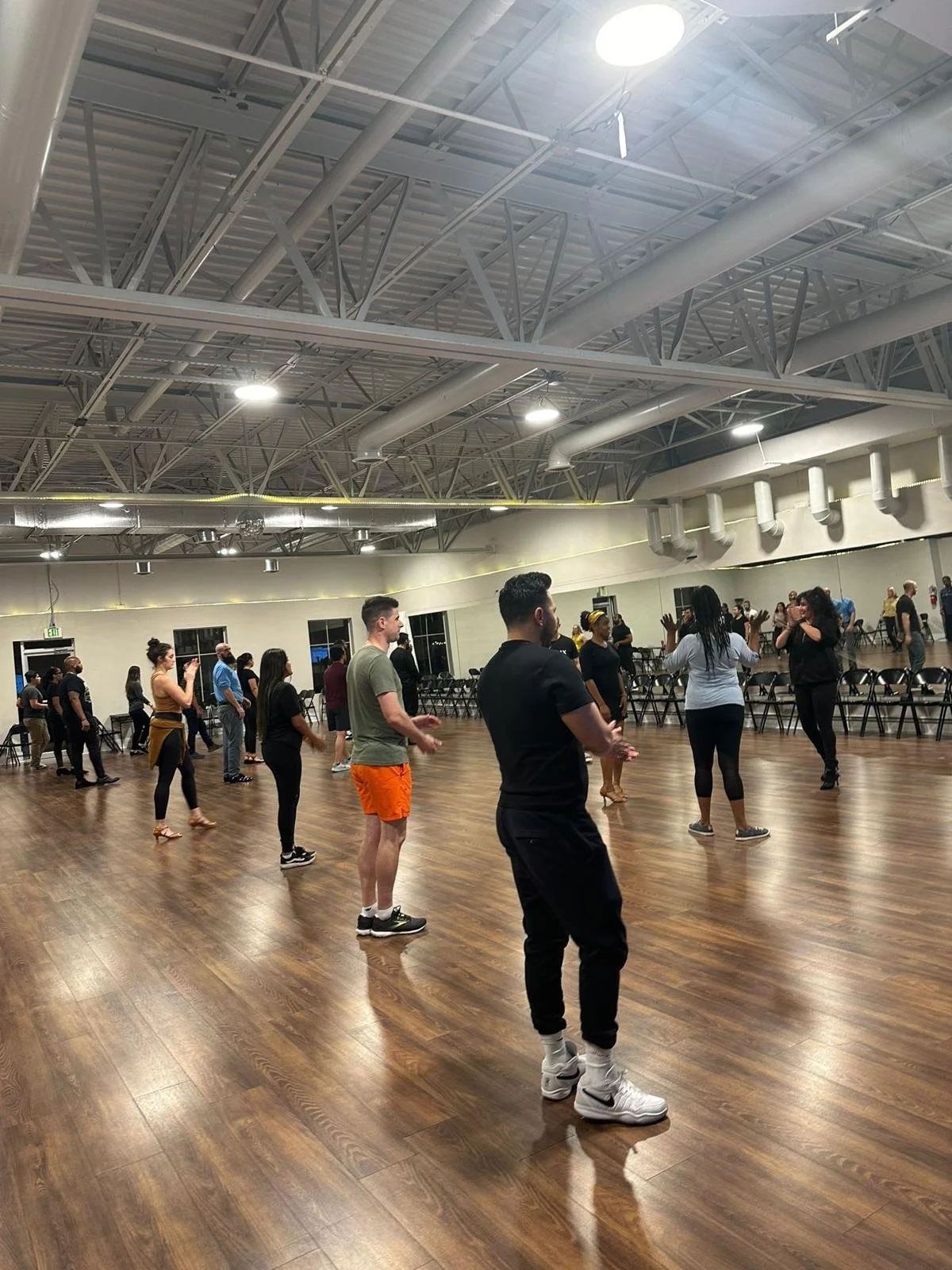 A diverse group of people practicing dance or exercise in a spacious, industrial-style studio with a high ceiling, exposed ductwork, and wooden floors. Some participants are standing in a semi-circle, following instructions from an instructor.