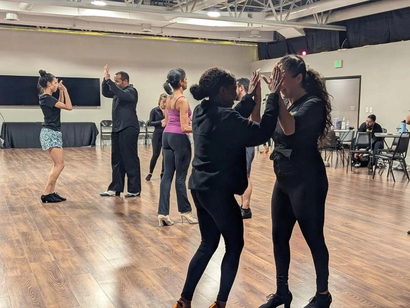 Two women dancing closely with their hands on each other's face in the foreground, other couples dancing in the background in a dance studio with wood floor and black curtains.
