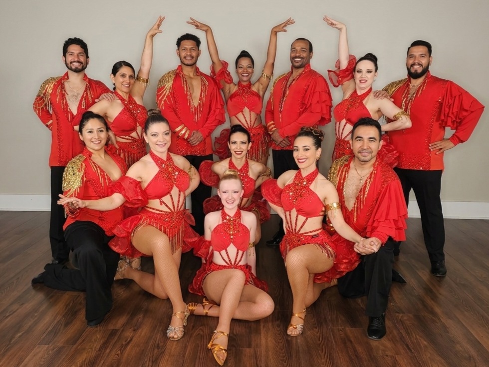 A group of twelve dancers in red and gold costumes posing together indoors, some standing and some kneeling, with a plain gray wall in the background.