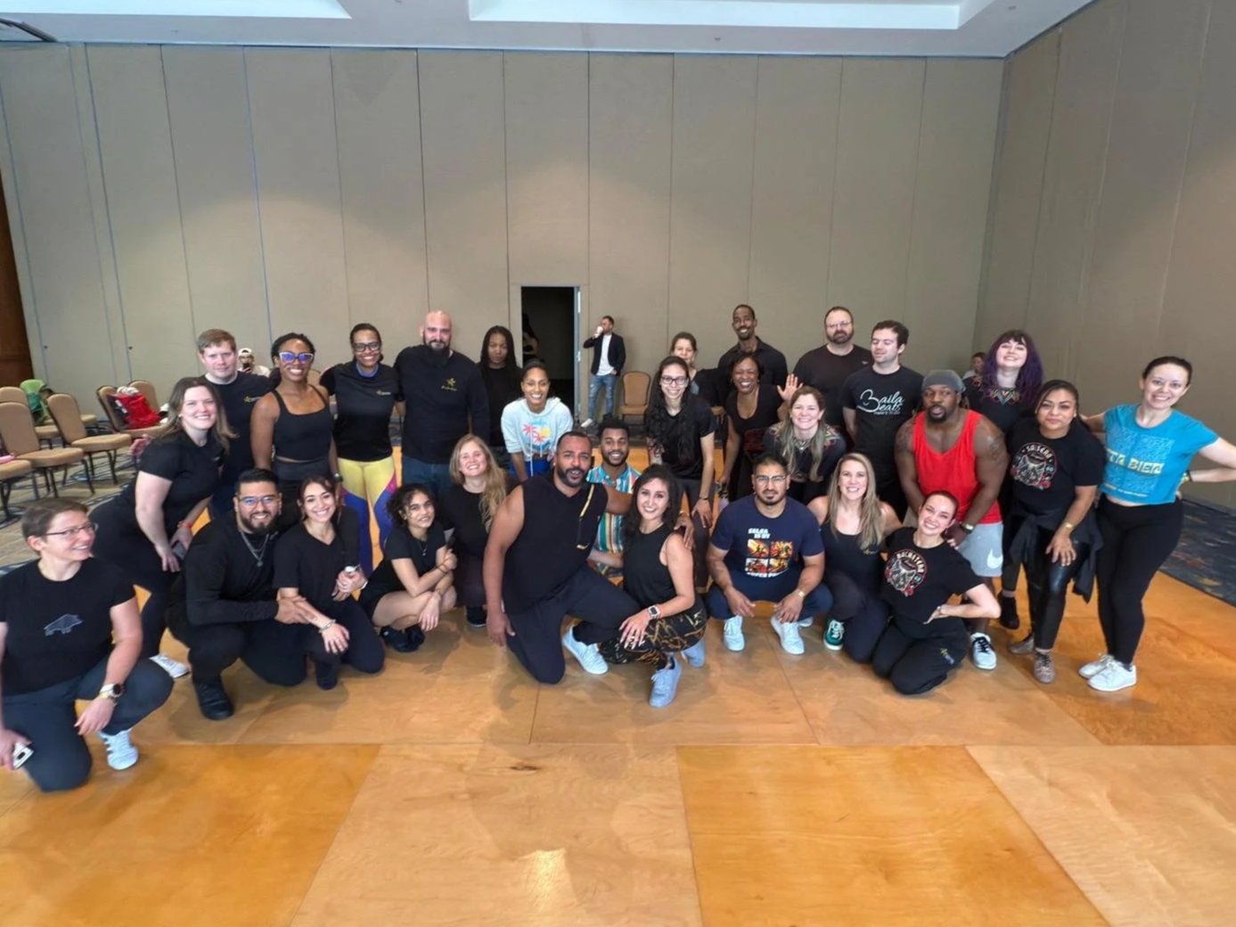 A large group of diverse people posing for a group photo in a spacious room with beige walls and wooden flooring. They appear happy and cheerful.