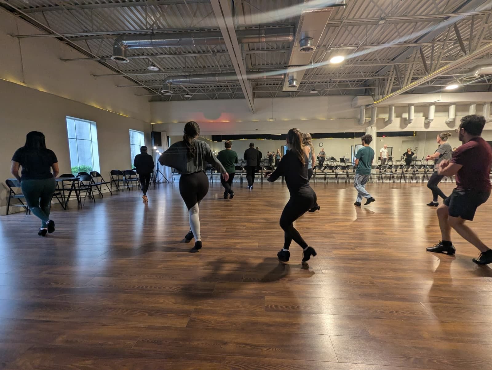 People practicing or dancing in a large studio with wooden floors and mirrors at the back. The studio is well-lit with natural light coming through large windows.