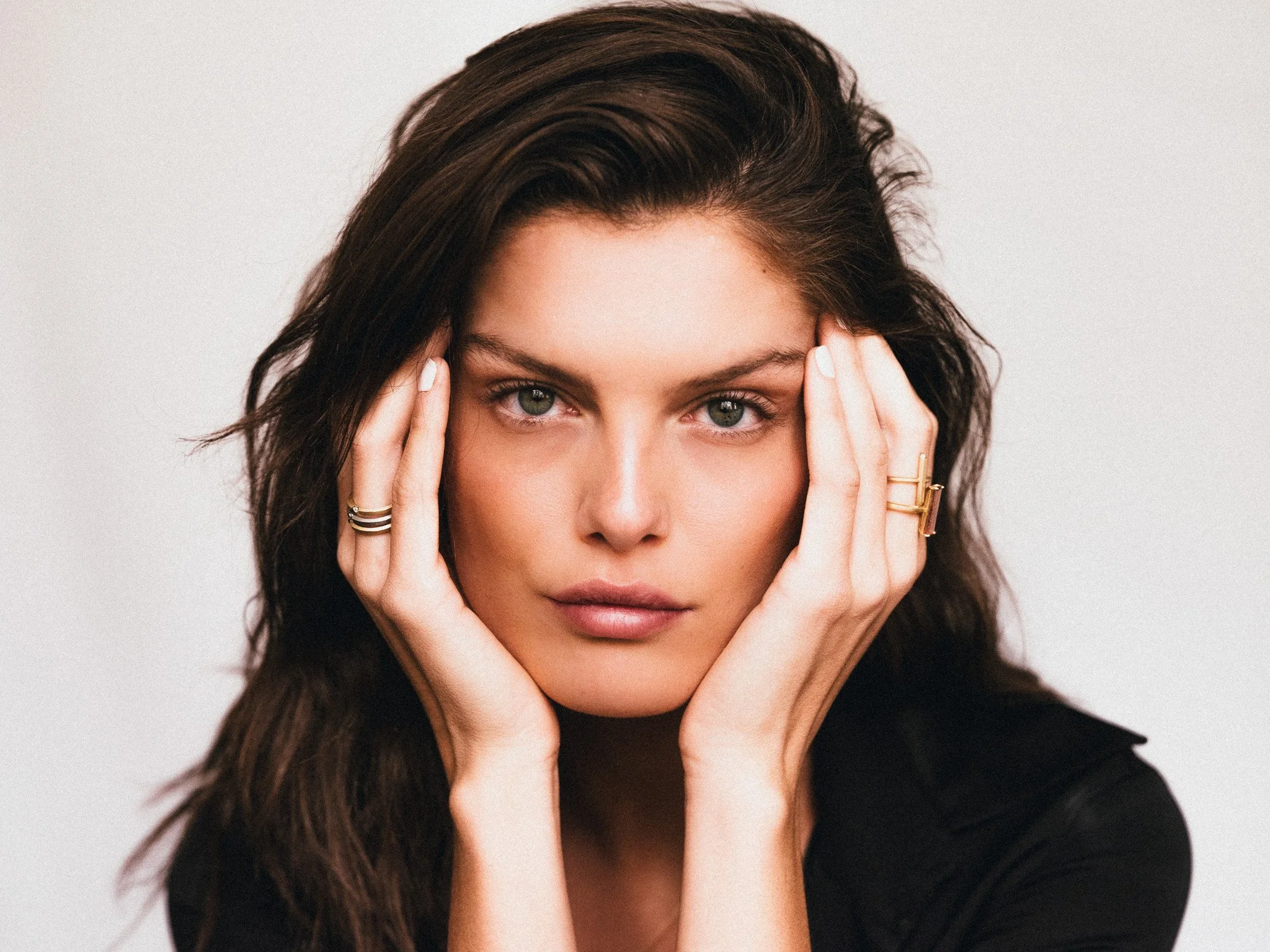 Close-up portrait of a woman with long brown hair, wearing rings and a black top, holding her face with both hands against a plain white background.