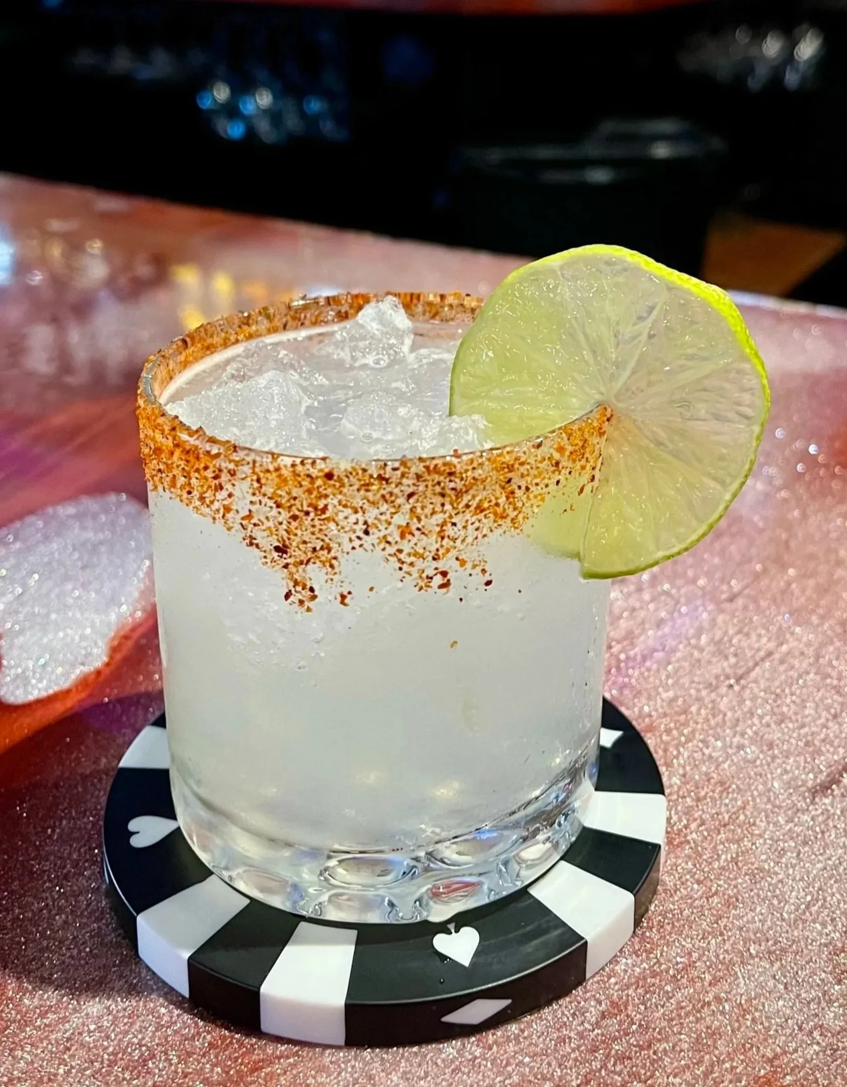 A glass of margarita with a salted rim and a lime wedge, placed on a black and white coasters with hearts, on a sparkly pink surface.