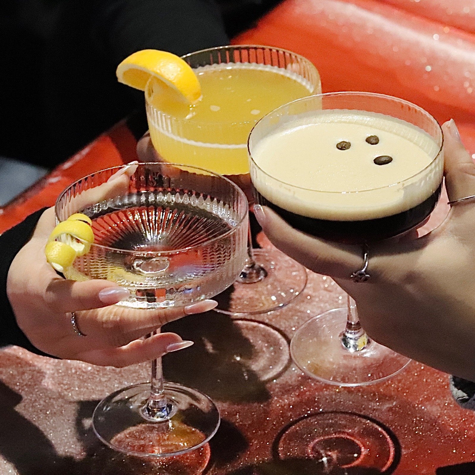 Three cocktails being clinked in glasses, one with a lemon wedge, on a table with a red textured surface.