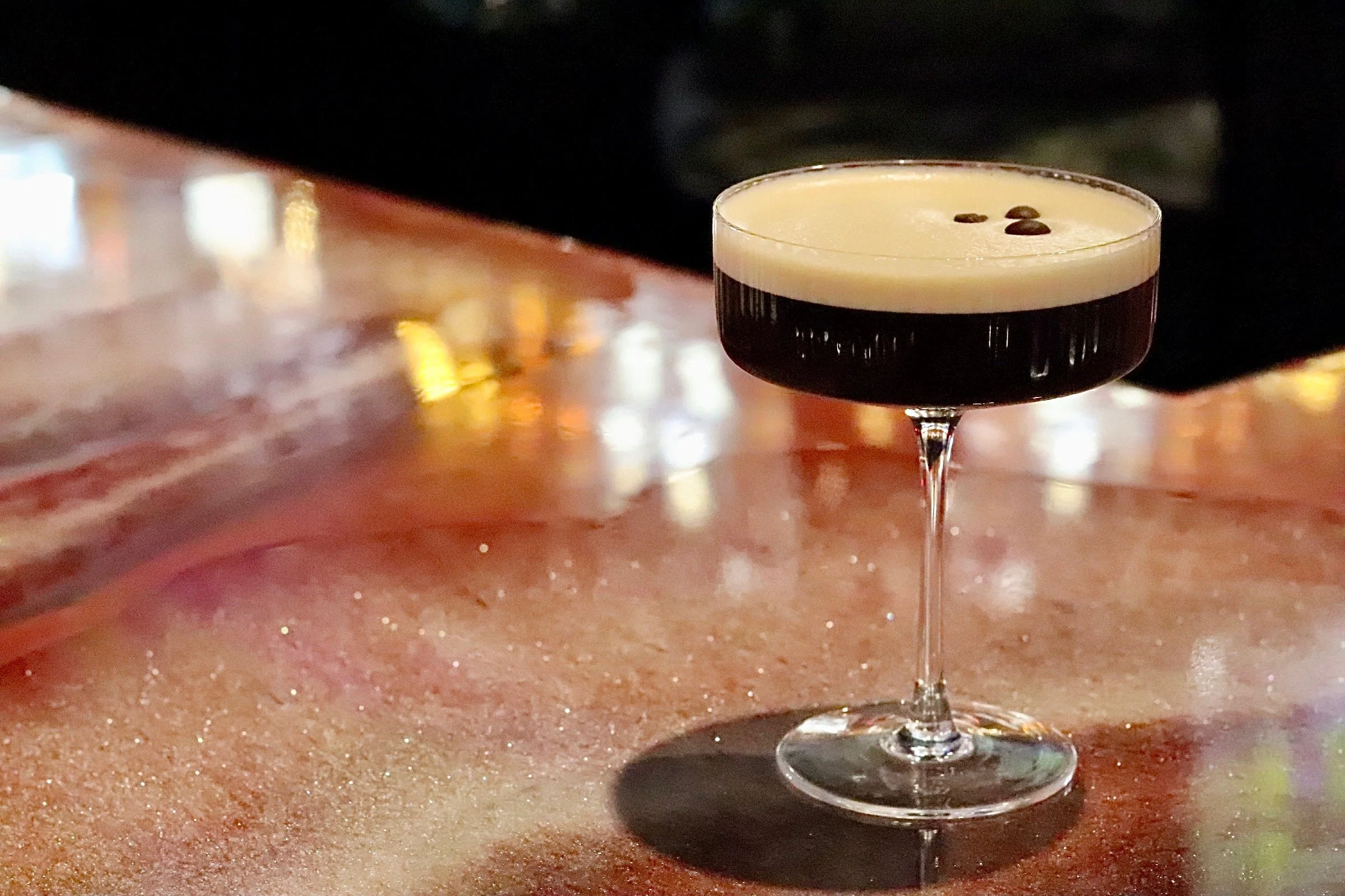 A cocktail glass with a dark layered drink topped with foam and coffee beans on a polished bar counter.