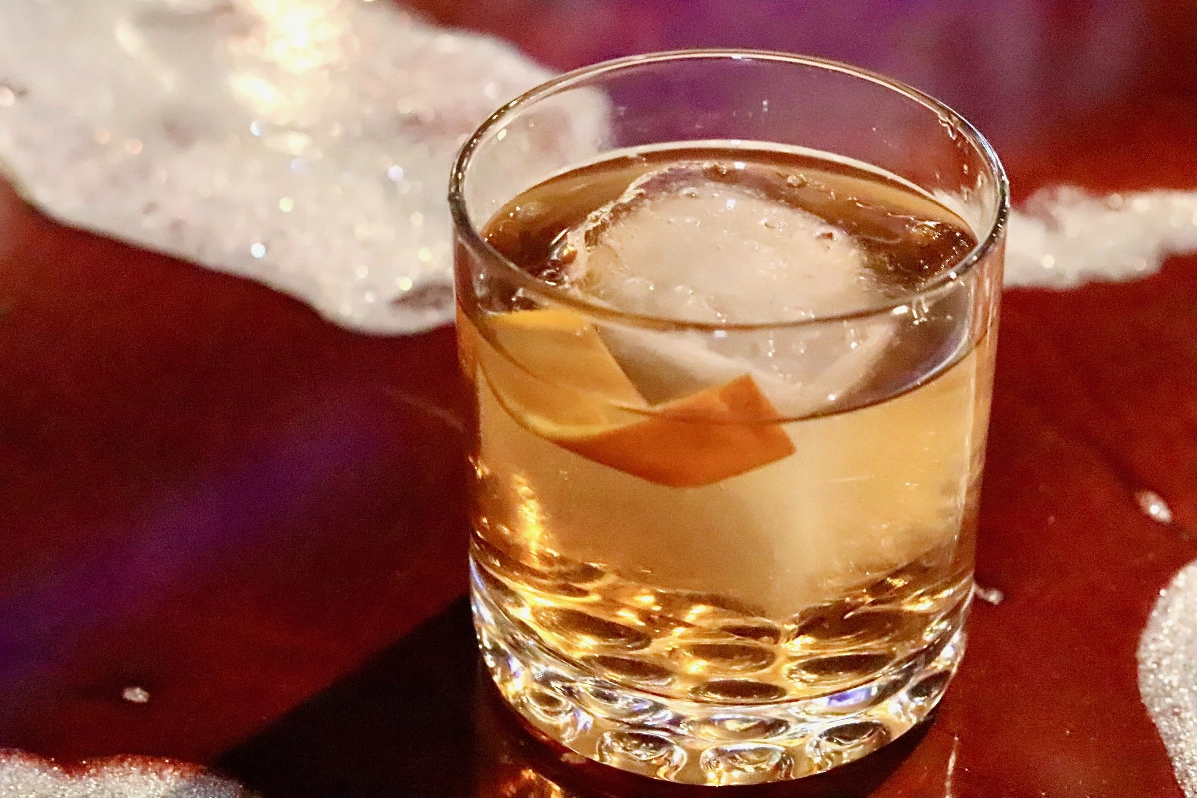 A glass of whiskey with a large ice cube and a twist of lemon on a dark wooden surface with glittering decorations around.