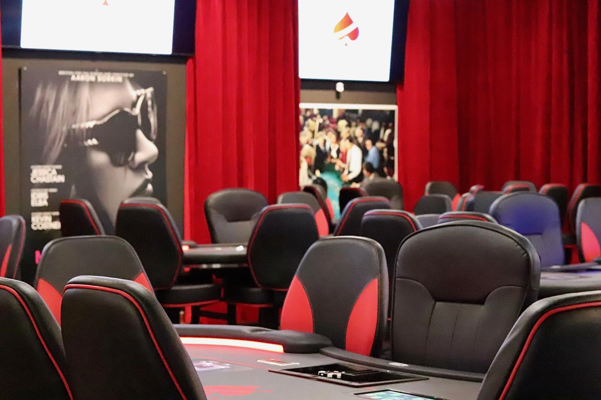 A conference room with black and red gaming chairs arranged around tables, red curtains, and large screens displaying promotional images.