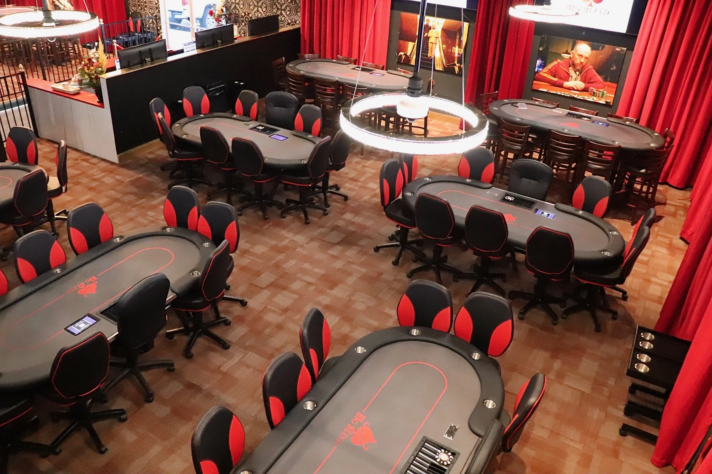 An empty poker room with multiple poker tables and chairs, large red curtains, and a TV screen on the wall showing a man playing poker.
