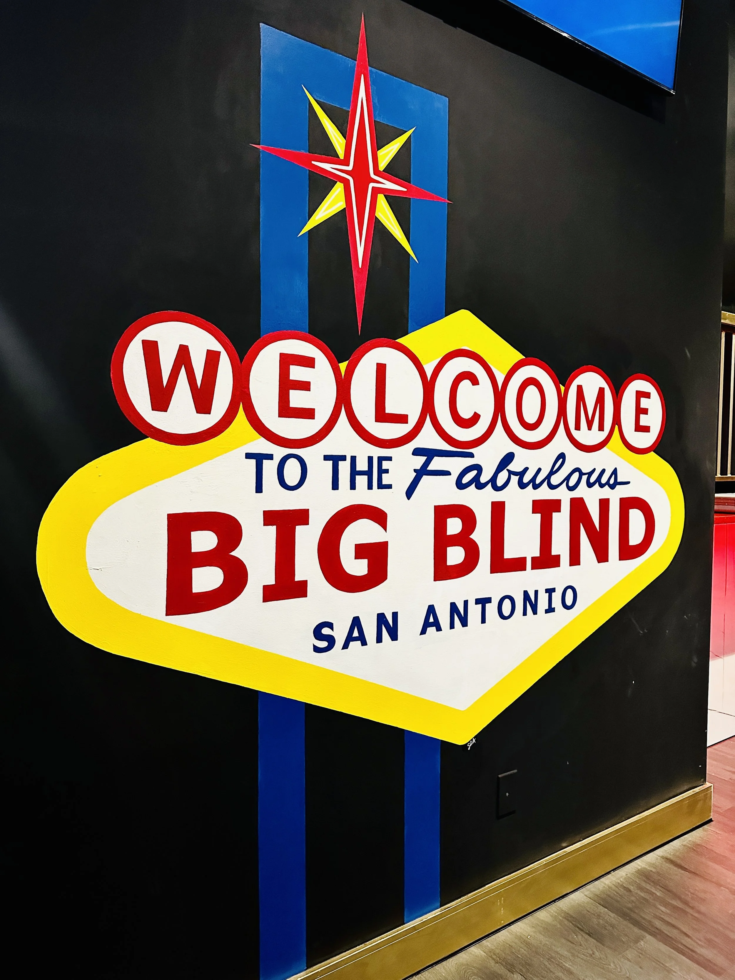 Colorful sign reading 'Welcome to the Fabulous Big Blind San Antonio' with a star graphic at the top, mounted on a black wall with blue stripes, in an indoor setting.