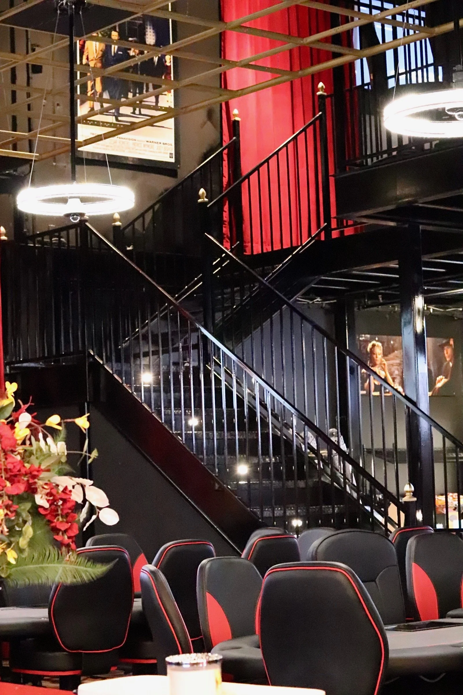 Interior of a restaurant or event space with black and red seating, a staircase with black railings, and large circular light fixtures hanging from the ceiling. There are framed posters and artwork on the walls and a floral arrangement in the foreground.