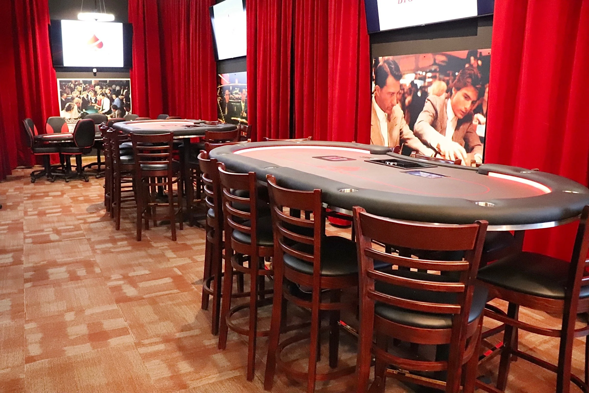 A poker room with a large oval poker table surrounded by chairs, red curtains, and large screens displaying images of people in a casino setting.