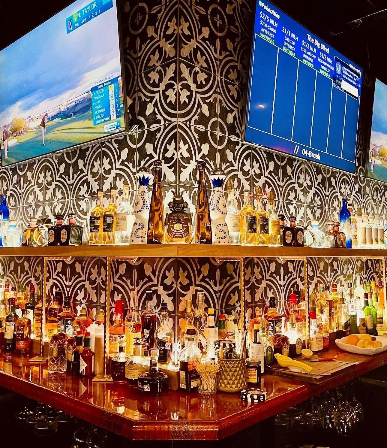 Bar counter with various liquor bottles, decorative vases, and glasses, with patterned tile wall and two television screens above showing golf scores.