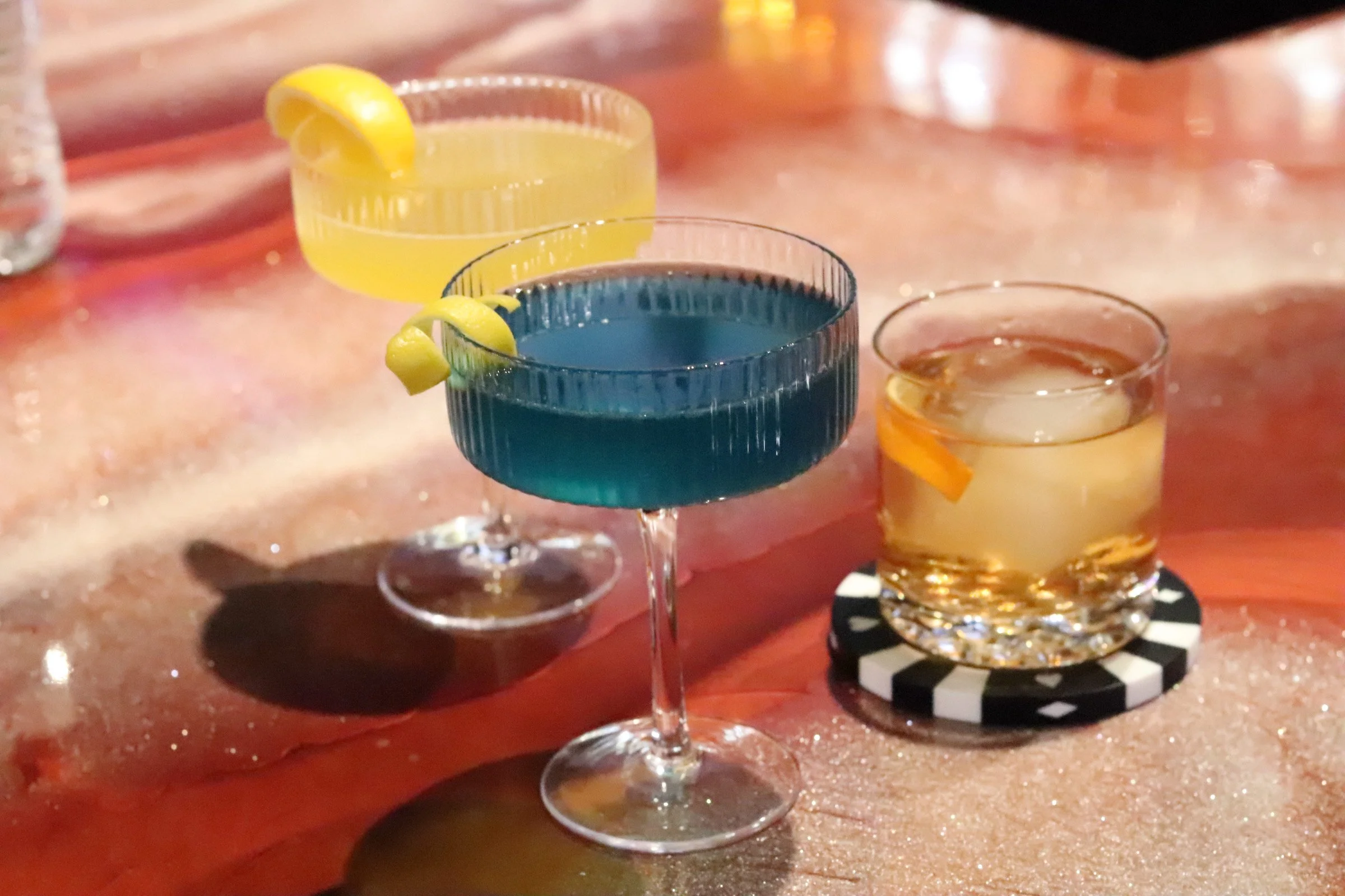 Three colorful cocktails on a wooden bar counter, each garnished with lemon or orange peel, with a blurred background.