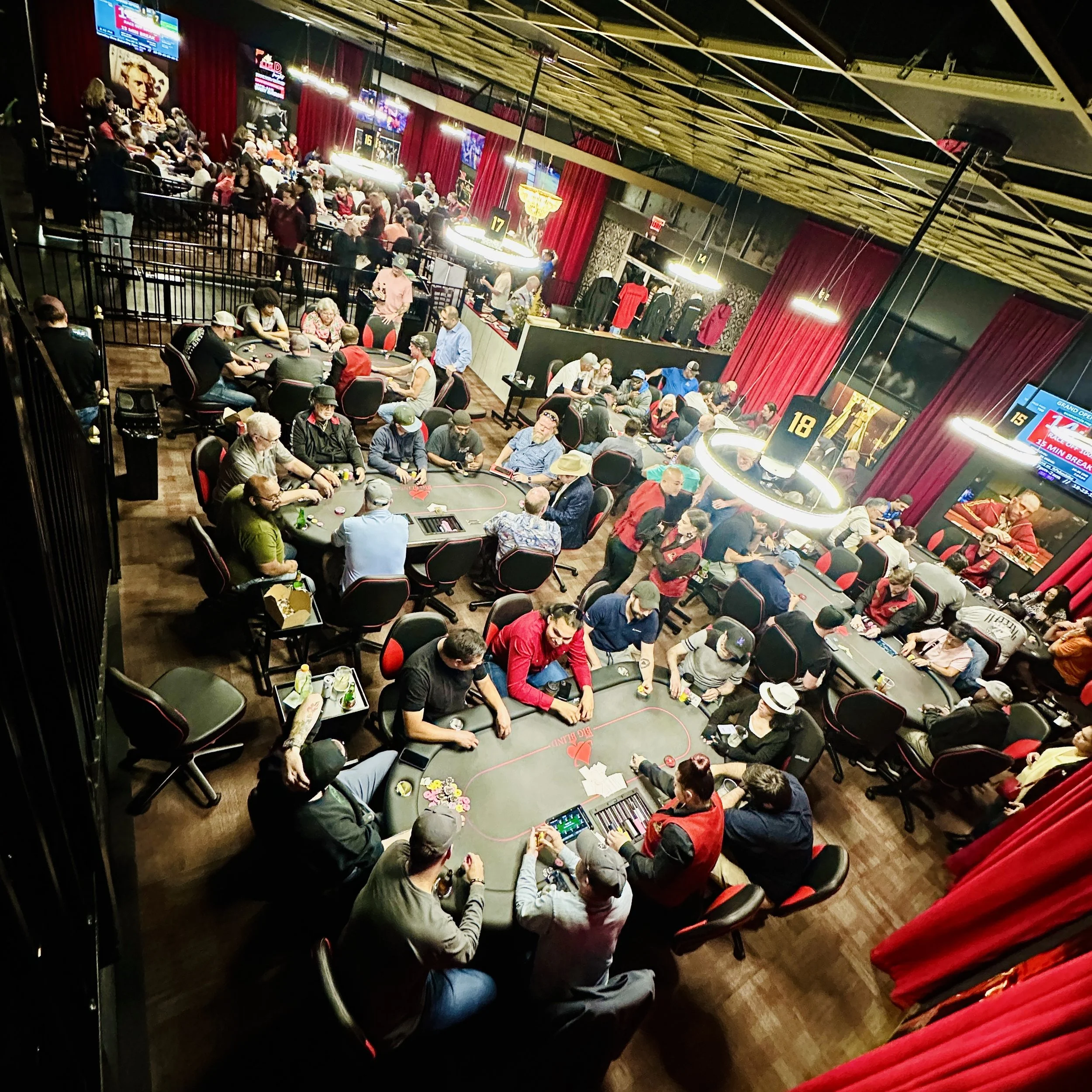 A large indoor poker tournament with multiple tables filled with players, some standing and some seated, under bright circular hanging lights. Red curtains and wall decorations, including vintage posters, are visible in the background. There are screens displaying tournament information and possible advertisements.