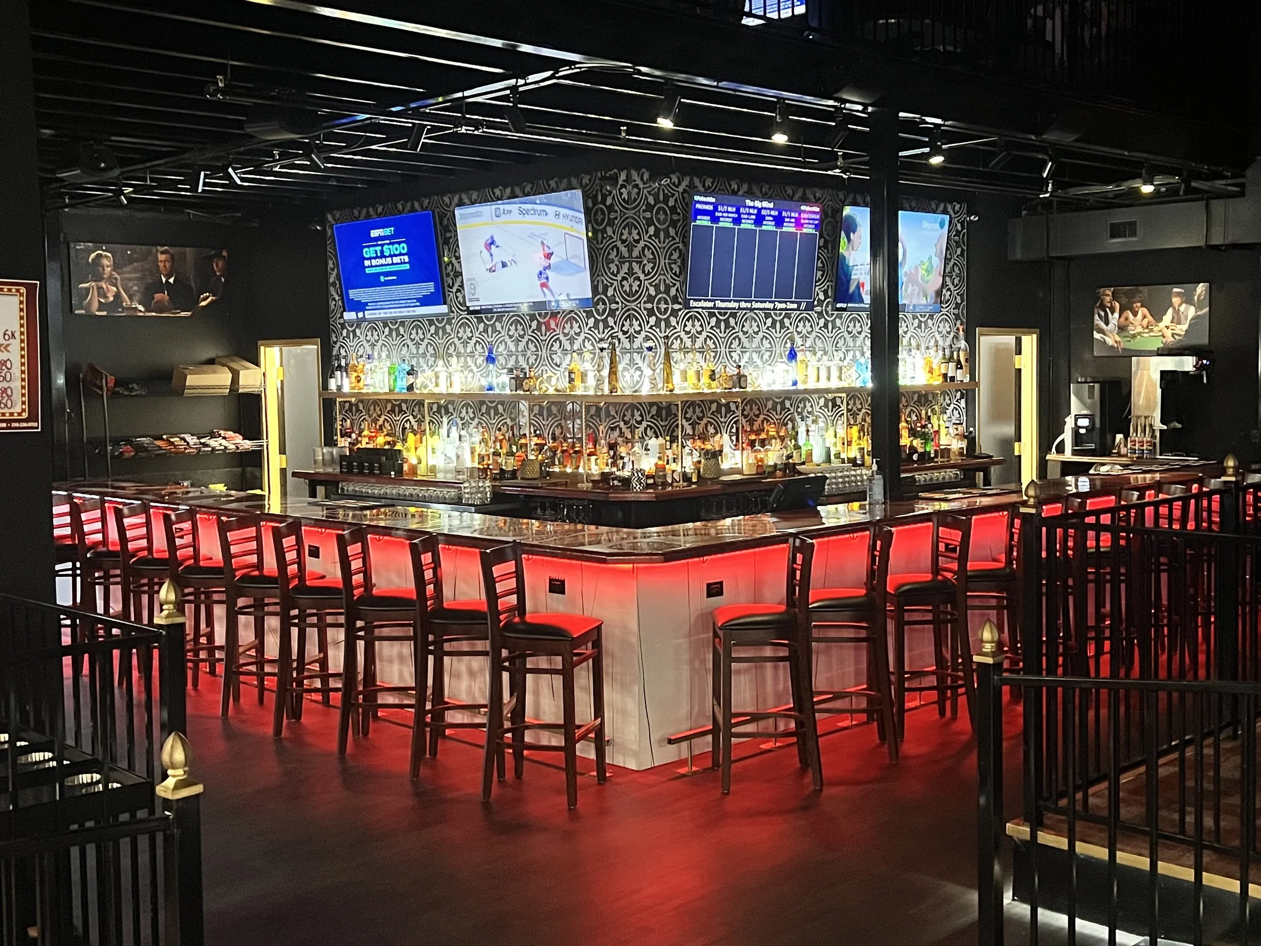 Empty bar with red glowing lights, high chairs, and multiple television screens displaying sports and advertisements in a dimly lit setting.