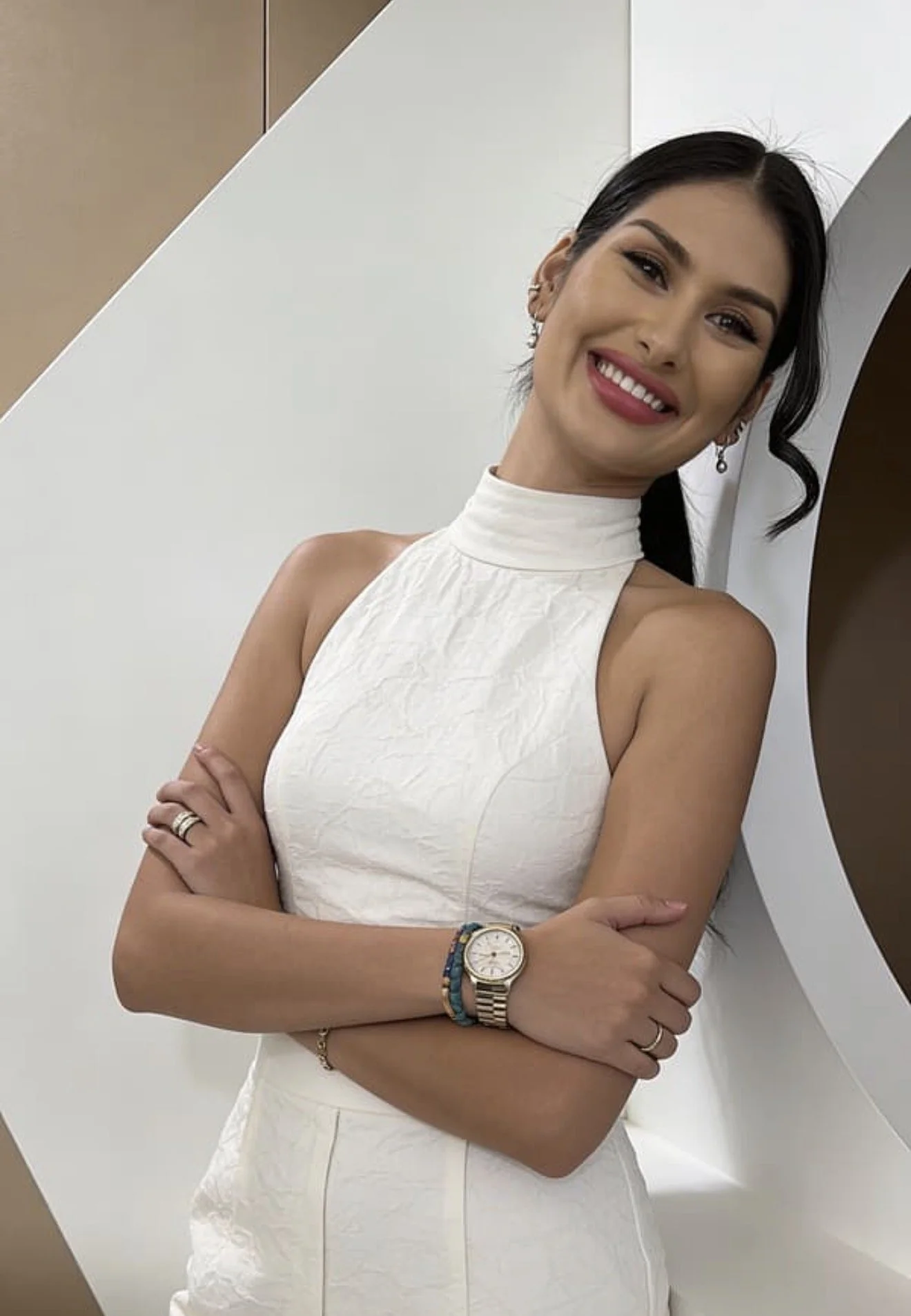 A woman smiling, with arms crossed, wearing a sleeveless white dress, a watch, and jewelry, standing against a modern, geometrical background.