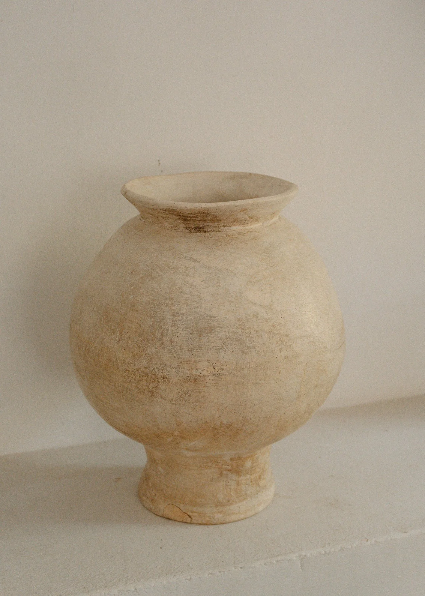 A beige ceramic vase with a rounded body, narrow neck, and flared opening, placed on a white surface against a plain off-white background.